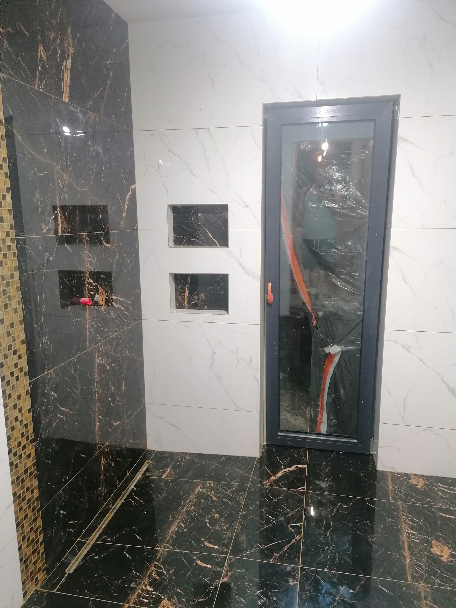 Wykończona łazienka z czarno-białymi płytkami imitującymi marmur, z wnękami w ścianie i szklanymi drzwiami prysznicowymi zabezpieczonymi folią ochronną.