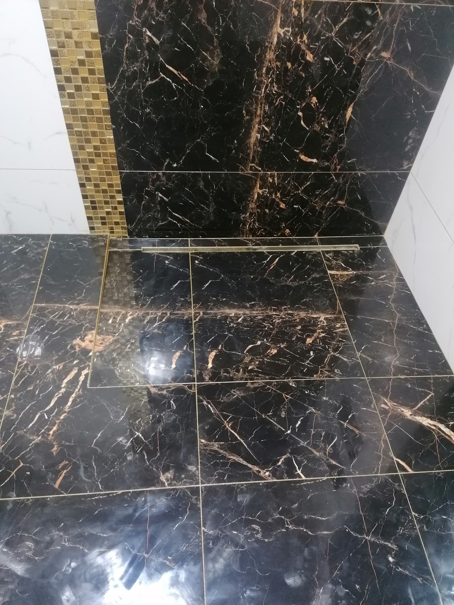 Wykończona podłoga w łazience z czarnych płytek imitujących marmur z złotymi żyłami, widoczny odpływ liniowy i fragment ściany z mozaiką w odcieniach złota i bieli.