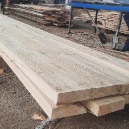 Stos świeżo przetartego, jasnego drewna na placu, z widoczną strukturą słojów i elementami maszyny do obróbki drewna w tle.