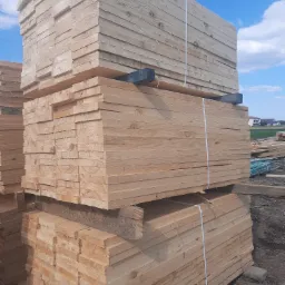 Ułożone w stosy świeże, jasne deski, zabezpieczone białymi taśmami, na tle błękitnego nieba z chmurami i fragmentu krajobrazu z domami.