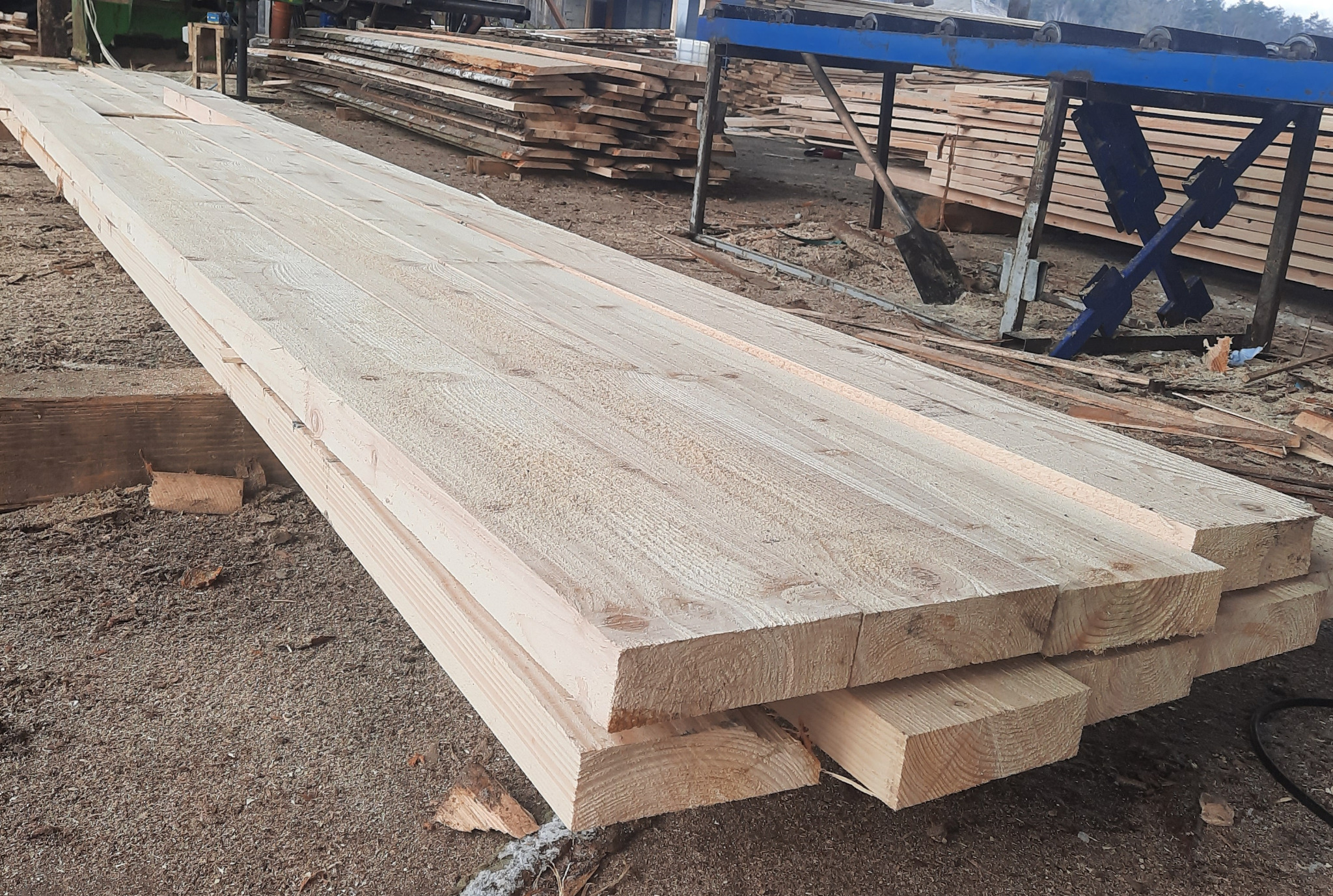 Stos świeżo przetartego, jasnego drewna na placu, z widoczną strukturą słojów i elementami maszyny do obróbki drewna w tle.