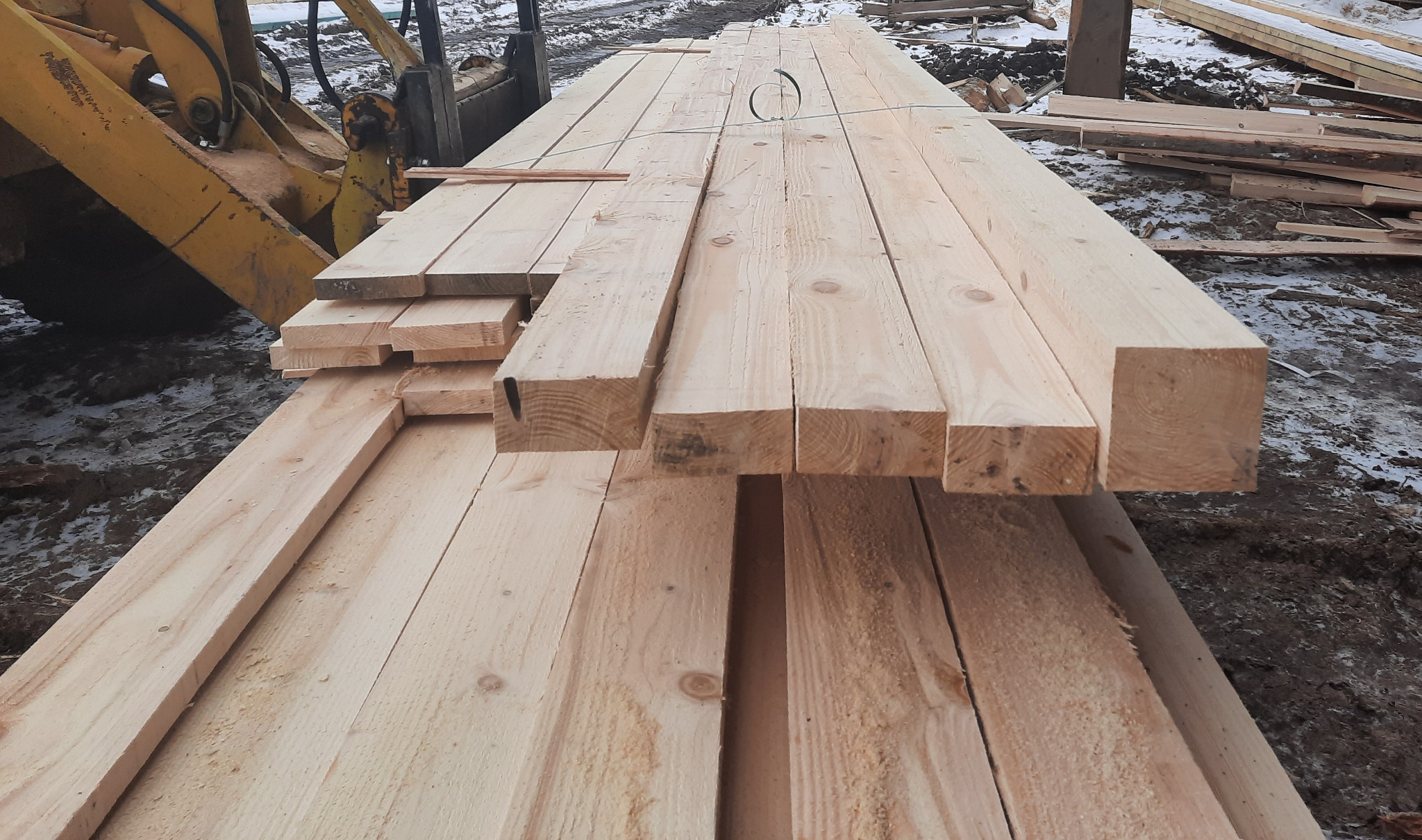 Świeżo przetarte, jasne drewno konstrukcyjne ułożone w stos, z widocznymi sękami i strukturą drewna, obok żółtej maszyny budowlanej na placu z resztkami śniegu.