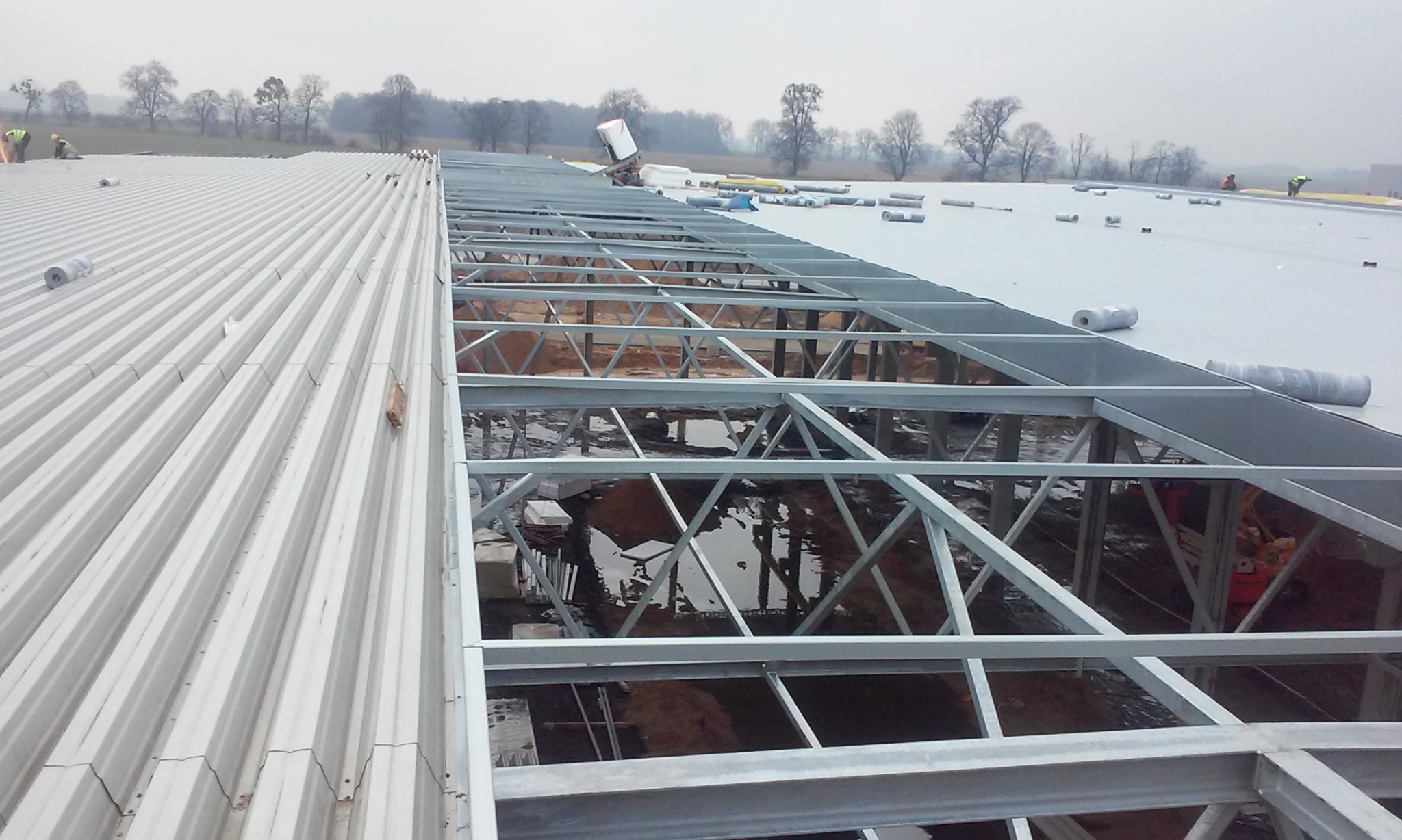 Widok z góry na konstrukcję stalową hali w trakcie budowy, z widocznymi rolkami materiału izolacyjnego i pracownikami na dachu w tle.