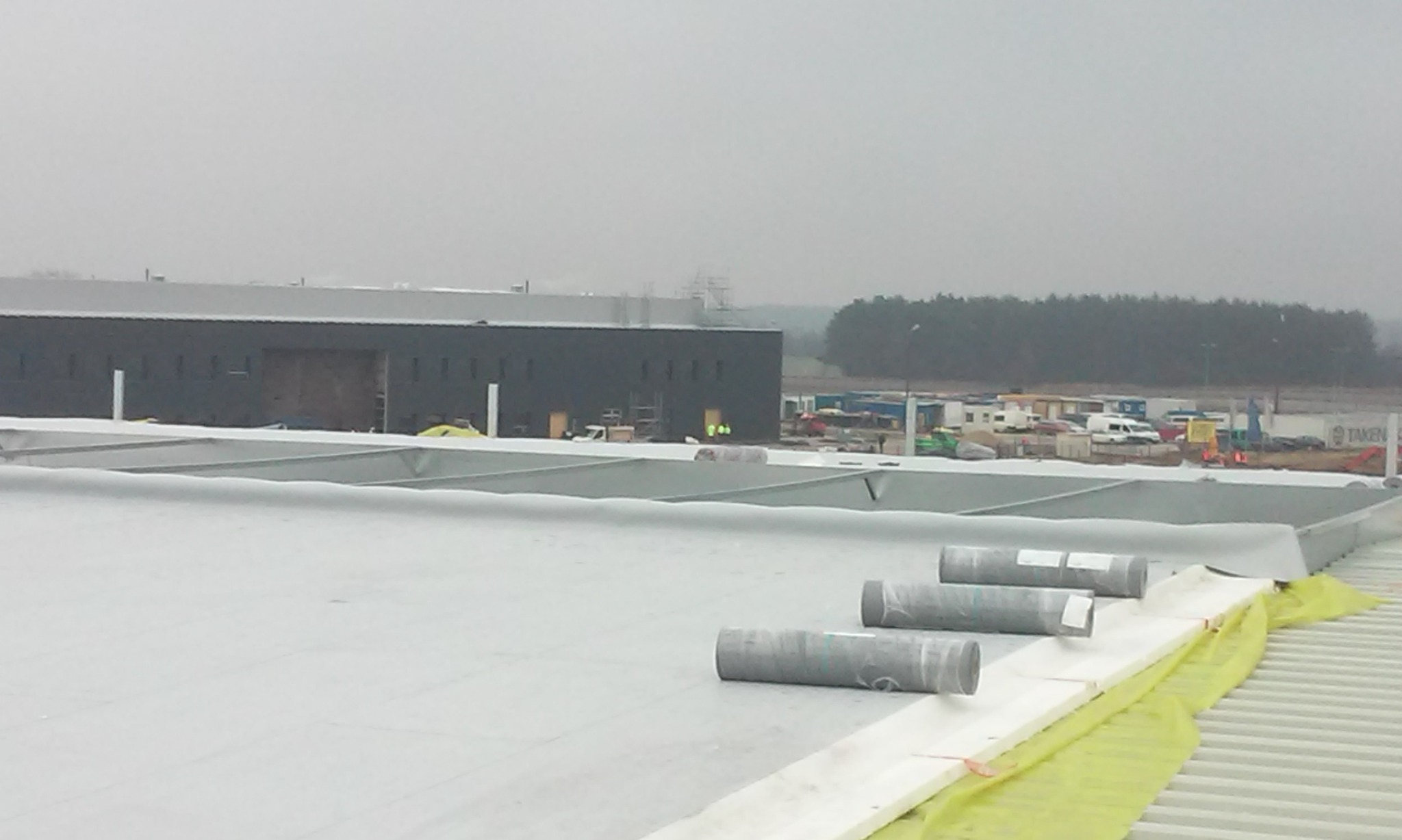 Widok z góry na dach hali w trakcie budowy z częściowo ułożonymi płytami warstwowymi, widoczne rolki materiału izolacyjnego i konstrukcja stalowa dachu na tle budynku przemysłowego i drzew.