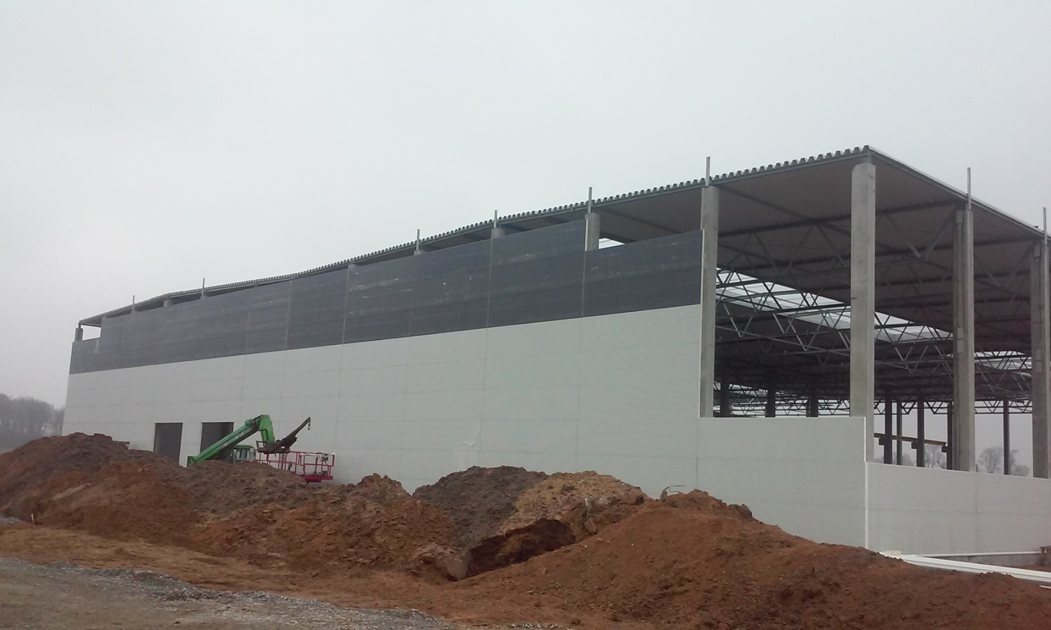 Wznoszenie hali produkcyjnej z widoczną konstrukcją stalową, częściowo obudowaną płytami warstwowymi, z użyciem podnośnika koszowego na tle hałd ziemi w pochmurny dzień.