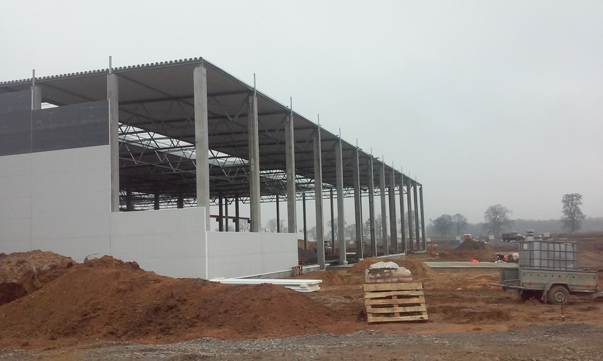 Budowa hali: szkielet konstrukcji stalowej z betonowymi słupami i częściowo zamontowanymi płytami warstwowymi, widoczne przygotowane fundamenty i teren budowy w pochmurny dzień.