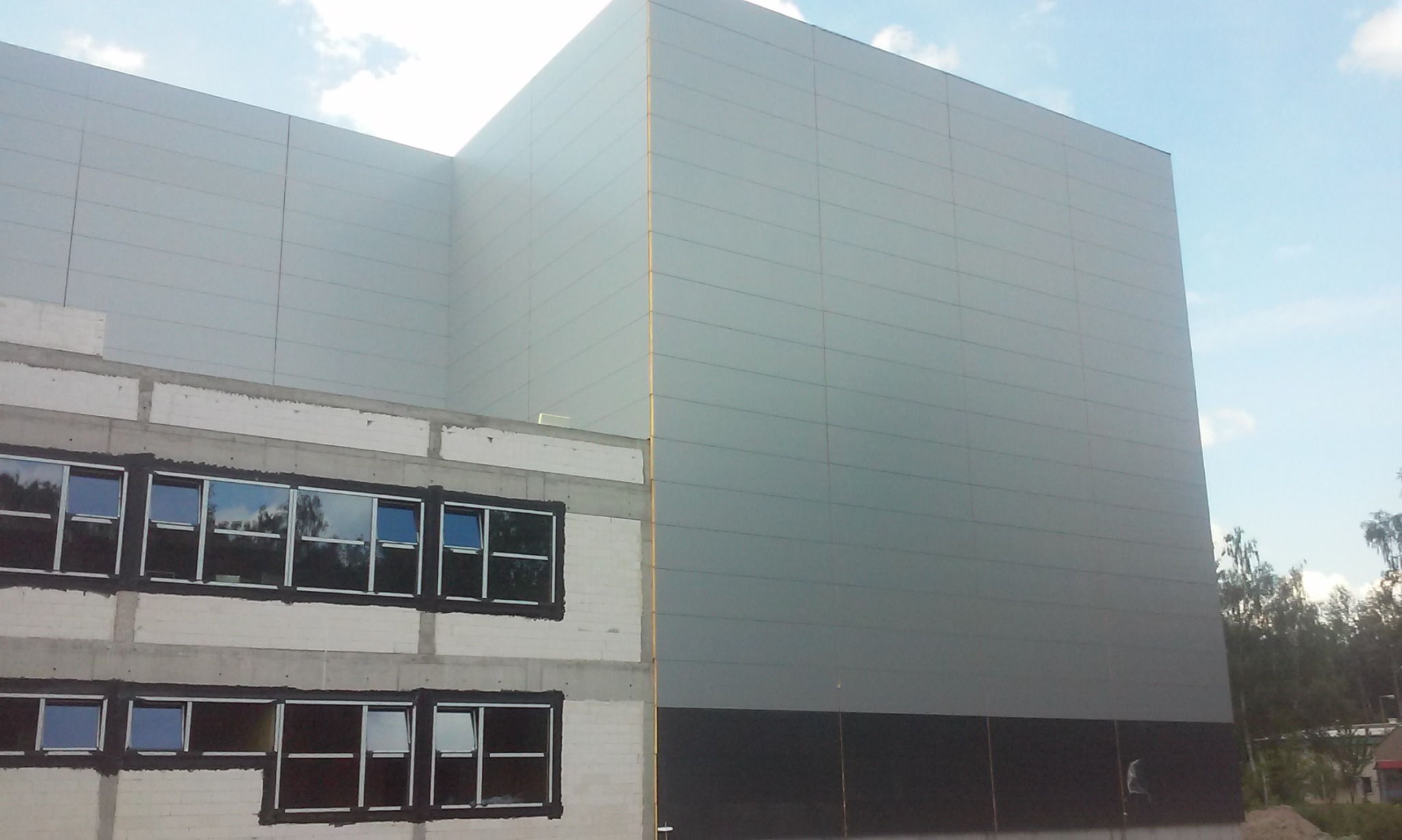 Wznoszenie budynku przemysłowego: fragment ściany z paneli warstwowych w odcieniach szarości, połączony z fragmentem ściany z betonu komórkowego z oknami w czarnych ramach, na tle błękitnego nieba.