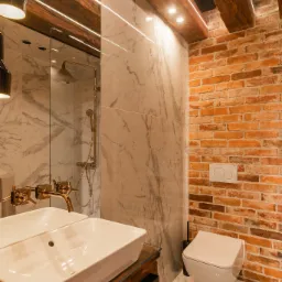 Nowoczesna łazienka z elementami loftu: biała umywalka nablatowa na drewnianym blacie, złota armatura, marmurowe płytki, ściana z czerwonej cegły, podwieszana toaleta i drewniane belki na suficie...