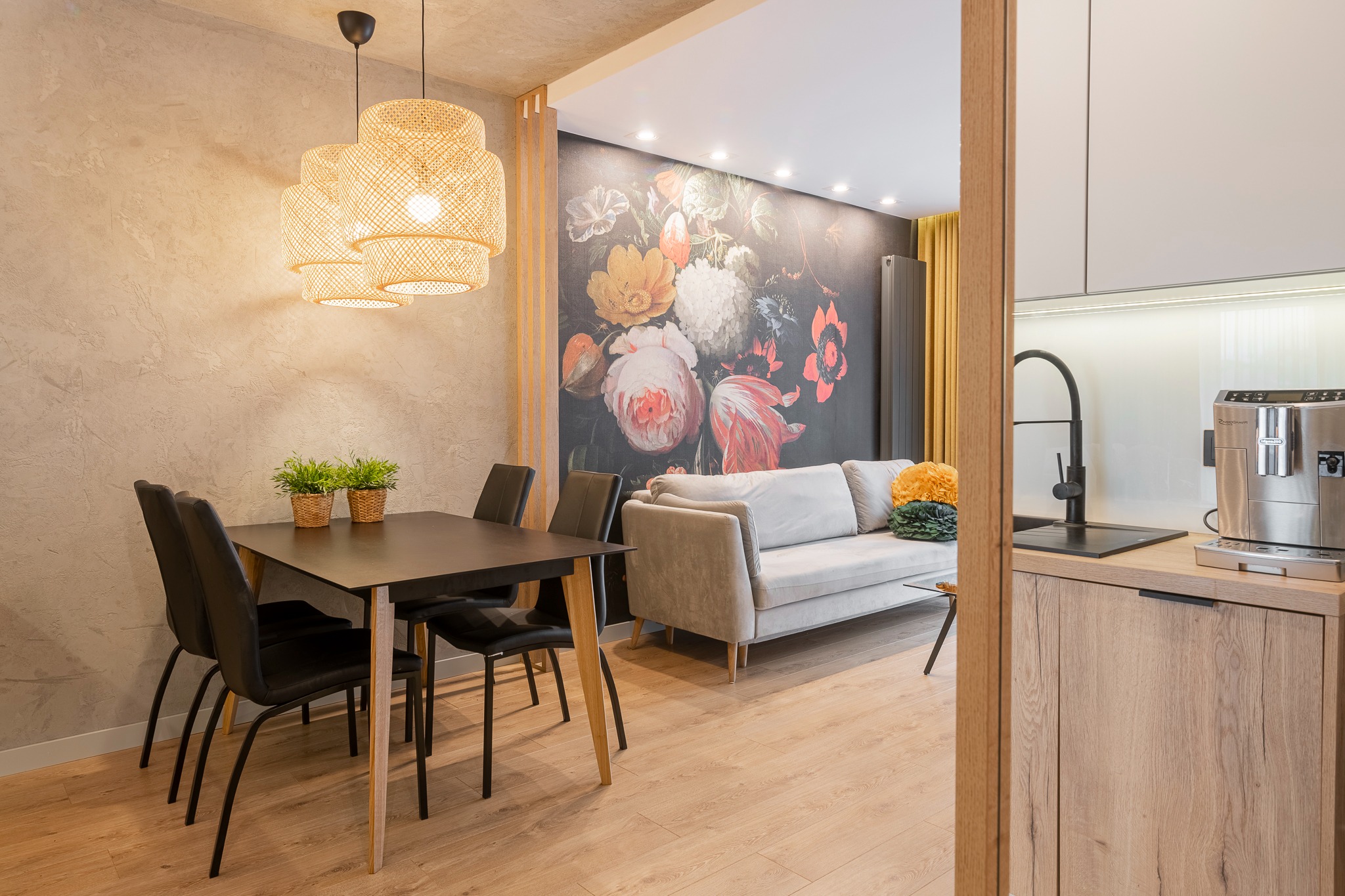 Nowoczesne wnętrze mieszkania z otwartą przestrzenią: jadalnia z czarnym stołem i lampami z wikliny, salon z szarą sofą i ścianą z motywem kwiatowym, oraz fragment kuchni z ekspresem do kawy.