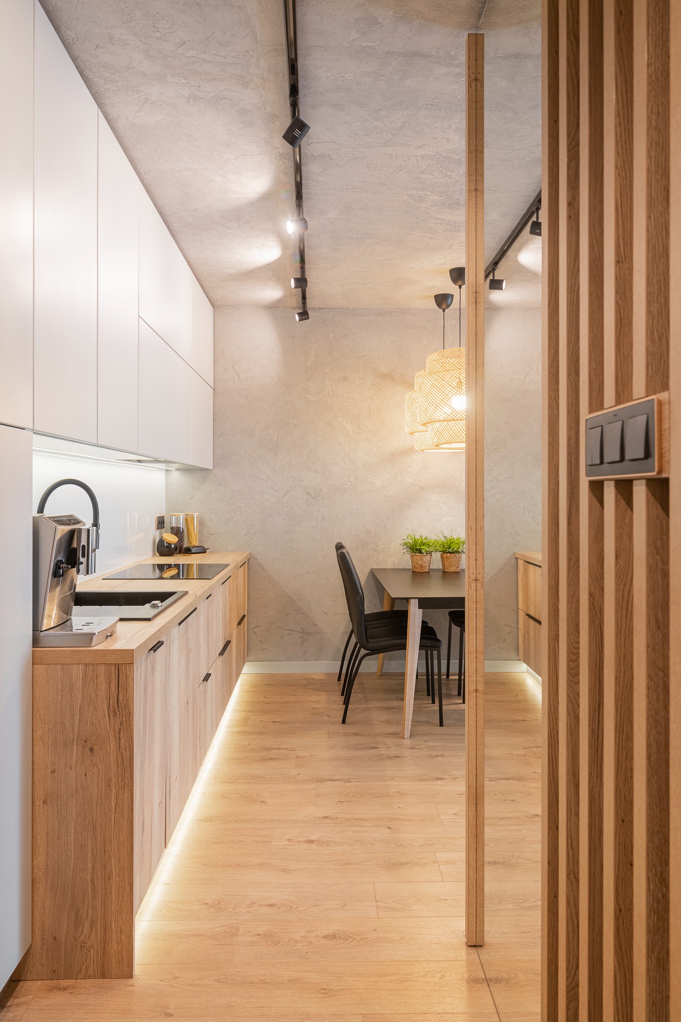 Nowoczesna kuchnia z drewnianymi frontami szafek, białymi górnymi szafkami, betonowym sufitem i oświetleniem punktowym, widok na jadalnię z drewnianą lampą i czarnym stołem z roślinami, oddzielona...
