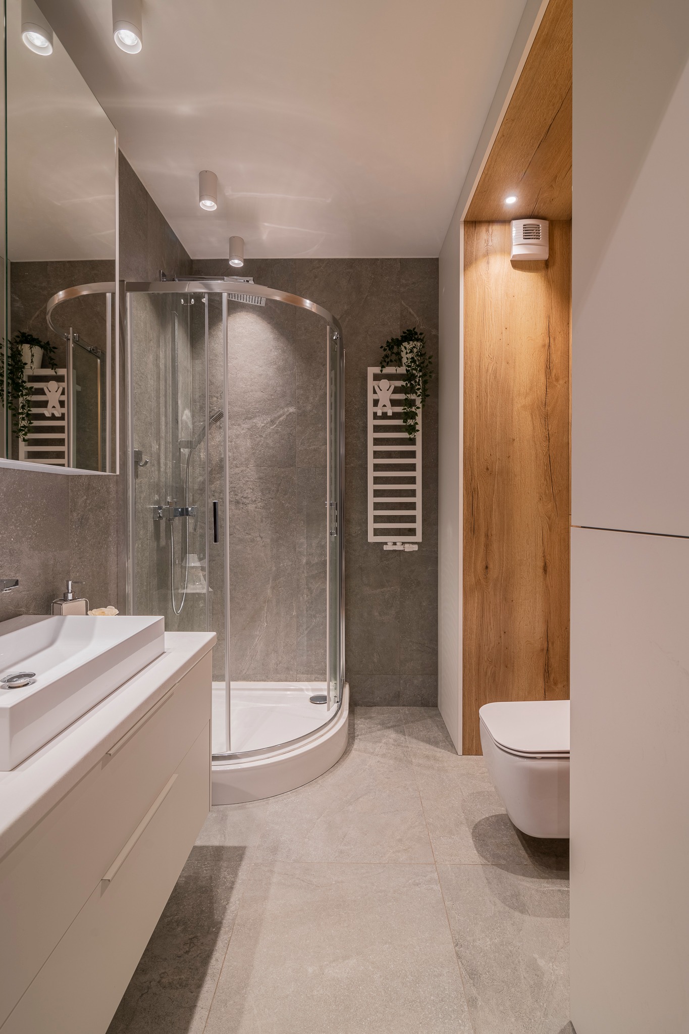 Nowoczesna łazienka z kabiną prysznicową, umywalką nablatową i toaletą podwieszaną, wykończona szarymi płytkami i elementami z drewna.