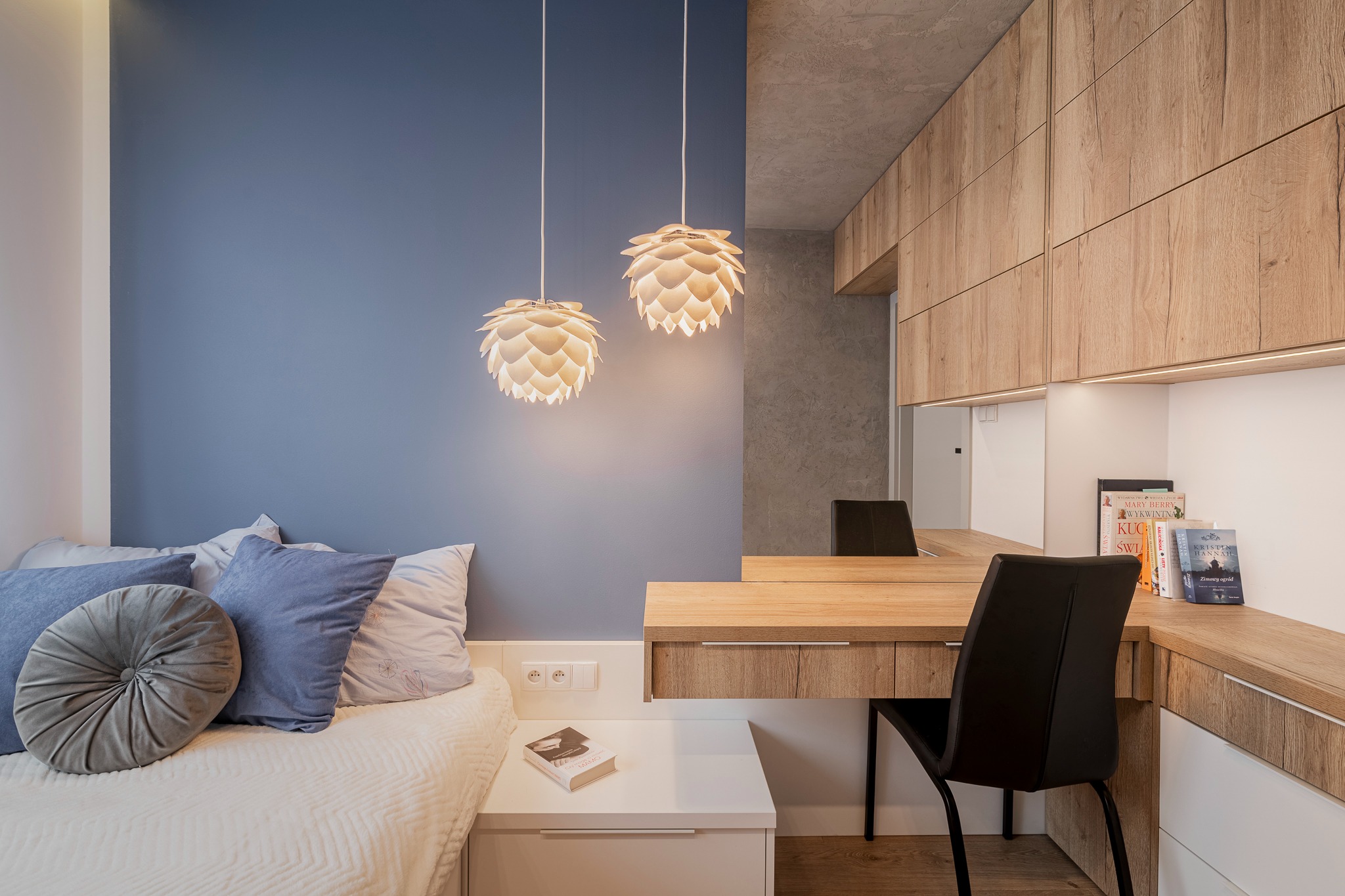 Nowoczesny pokój z niebieską ścianą, łóżkiem z dekoracyjnymi poduszkami, biurkiem z drewnianym blatem i designerskimi lampami wiszącymi w kształcie kwiatów.
