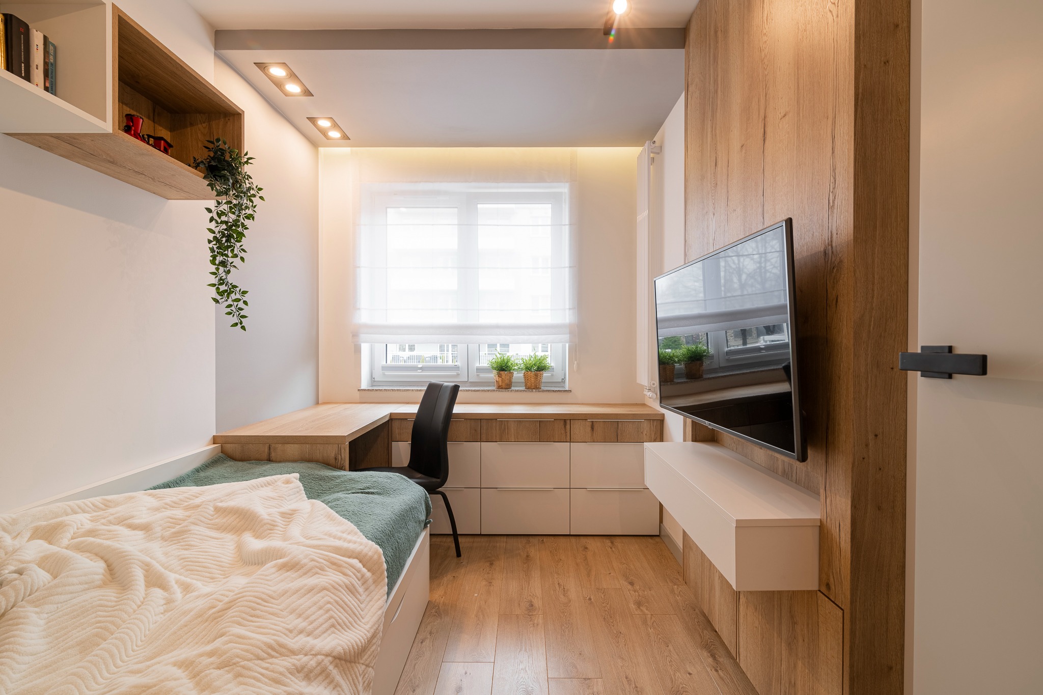 Funkcjonalny pokój młodzieżowy z łóżkiem, biurkiem pod oknem, telewizorem na drewnianej ścianie i wiszącą roślinką, oświetlony ciepłym światłem.