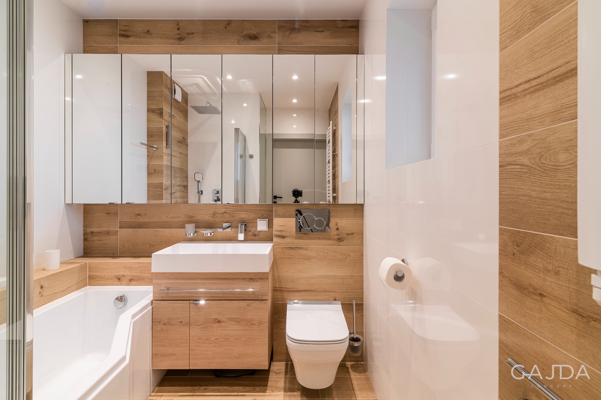 Nowoczesna łazienka z drewnopodobnymi płytkami, białą armaturą i lustrzaną szafką, widoczna wanna z parawanem nawannowym i toaleta.