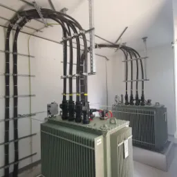 Dwa transformatory w pomieszczeniu z widocznymi kablami w czarnych peszlach, zamocowane do sufitu za pomocą metalowych konstrukcji wsporczych.