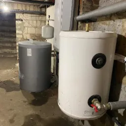 Dwa zbiorniki na wodę (biały i szary) w piwnicy o surowych ścianach, widoczne rury i zawory, podłoga lekko zabrudzona.