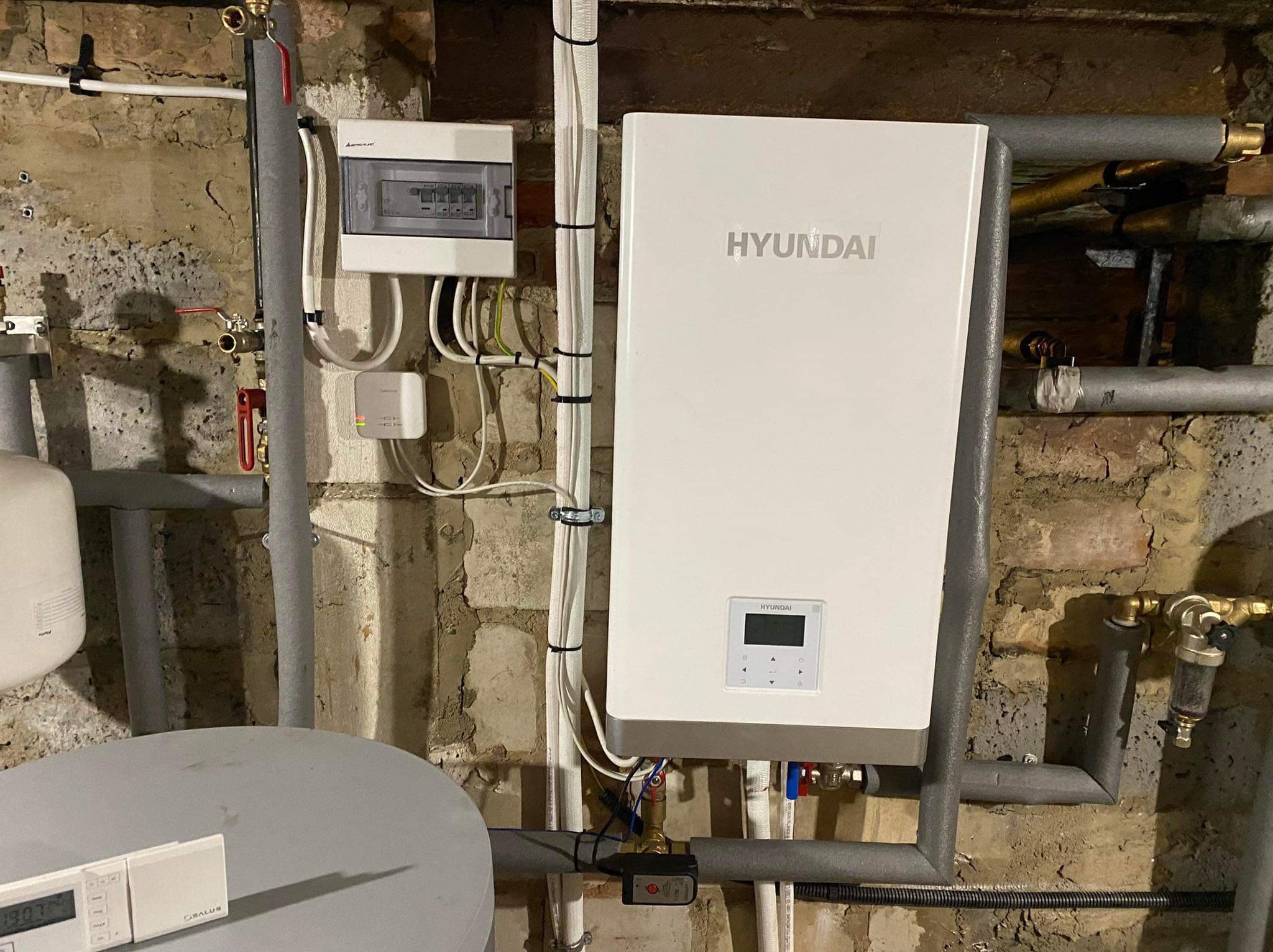 Biały piec grzewczy Hyundai zamontowany na ścianie z cegły, widoczne rury instalacji, skrzynka elektryczna i sterownik temperatury.