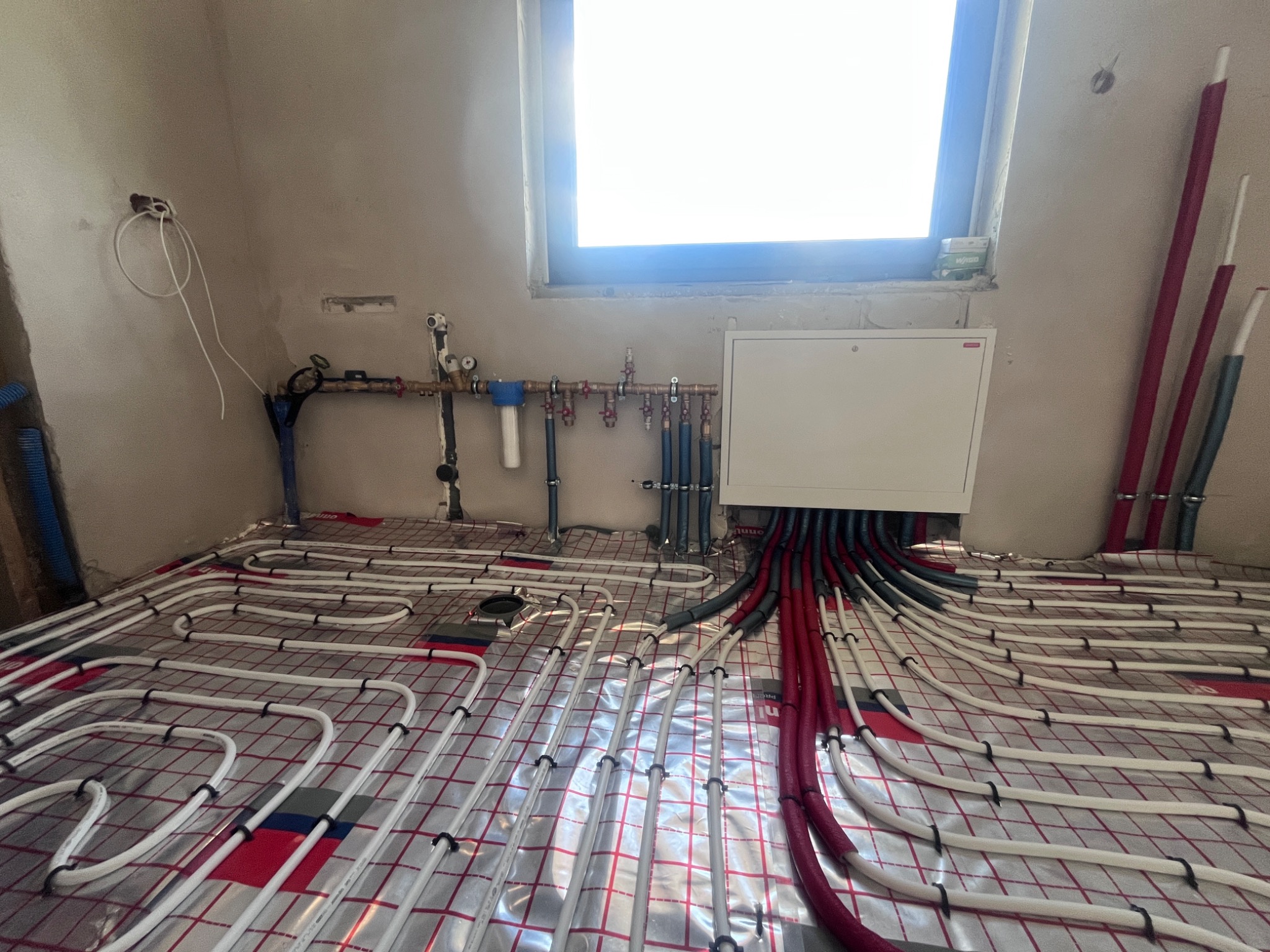 Białe rury ogrzewania podłogowego ułożone na folii z czerwoną kratą, rozdzielacz z rurami w kolorze szarym i czerwonym. W tle okno i instalacja wodna na ścianie.