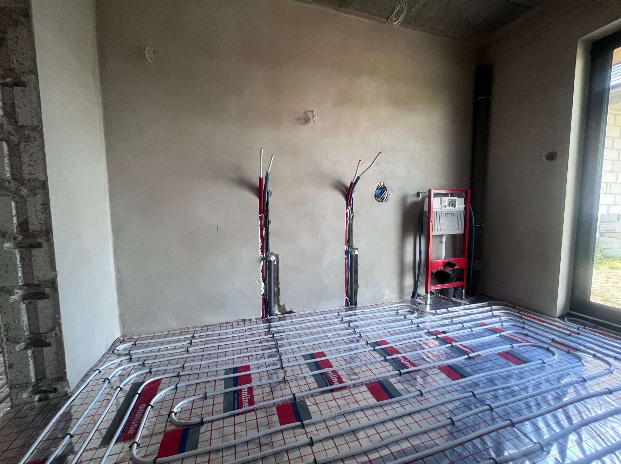 Układanie ogrzewania podłogowego w surowym wnętrzu. Widoczne rury na folii z nadrukiem, pionowe instalacje w ścianach i stelaż WC. Realizacja przed wylewką.