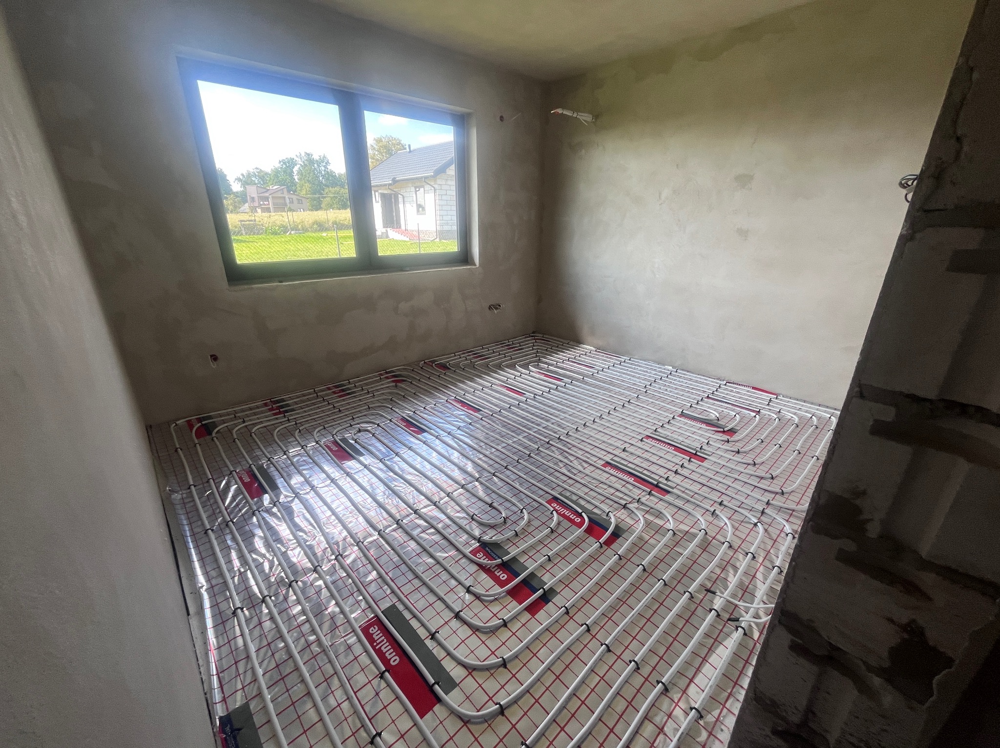 Białe rury ogrzewania podłogowego ułożone na folii w surowym pomieszczeniu z oknem. Widoczny fragment domu za oknem i surowe ściany.