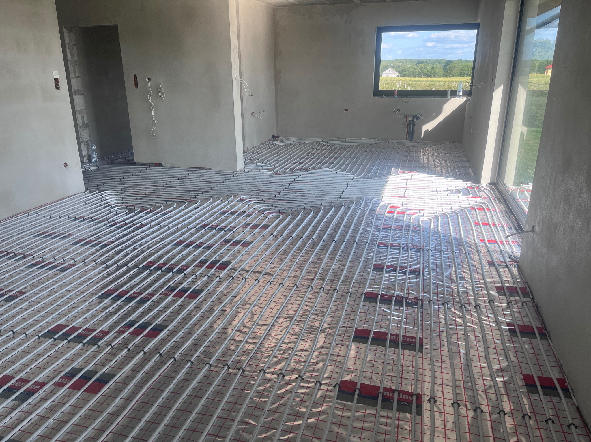 Białe rury ogrzewania podłogowego ułożone na folii w surowym wnętrzu z oknem i widokiem na zieleń. Etap instalacji przed wylewką.