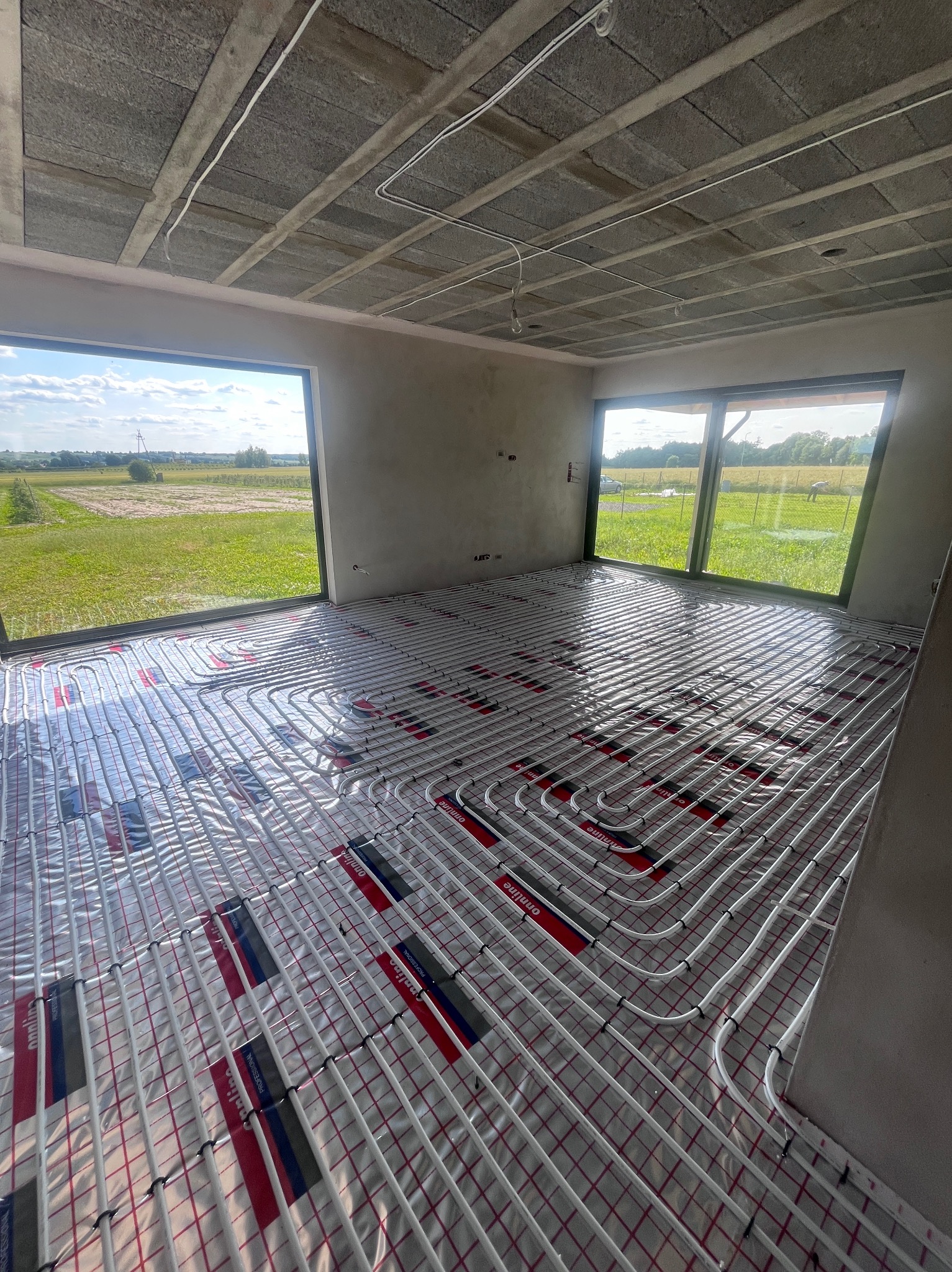 Białe rury ogrzewania podłogowego ułożone na folii z czerwoną siatką w jasnym pokoju z dużymi oknami i betonowym sufitem. Widok na zewnątrz.