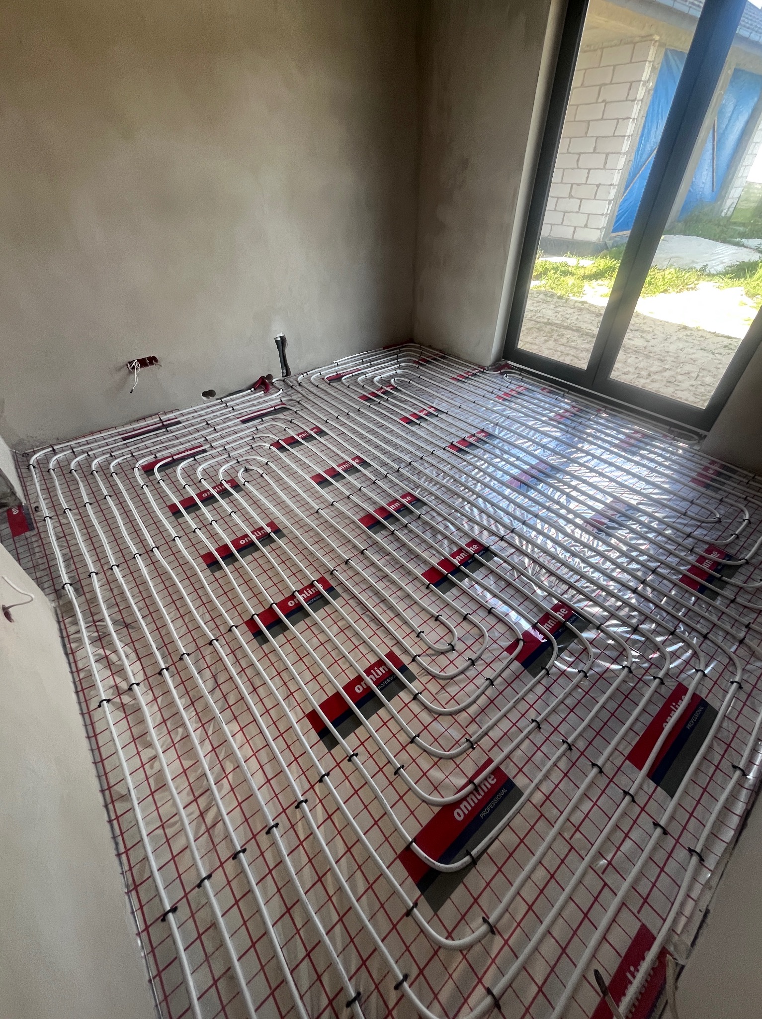 Białe rury ogrzewania podłogowego ułożone na folii z czerwoną siatką w surowym pomieszczeniu z oknem. Widoczne elementy mocujące i fragment instalacji na ścianie.