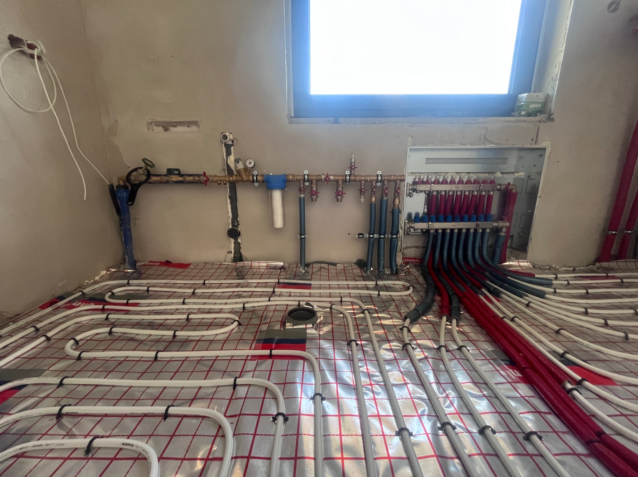 Rozłożone rury ogrzewania podłogowego na folii izolacyjnej, widoczny rozdzielacz z rurami w kolorze czerwonym i niebieskim. Instalacja w trakcie realizacji.