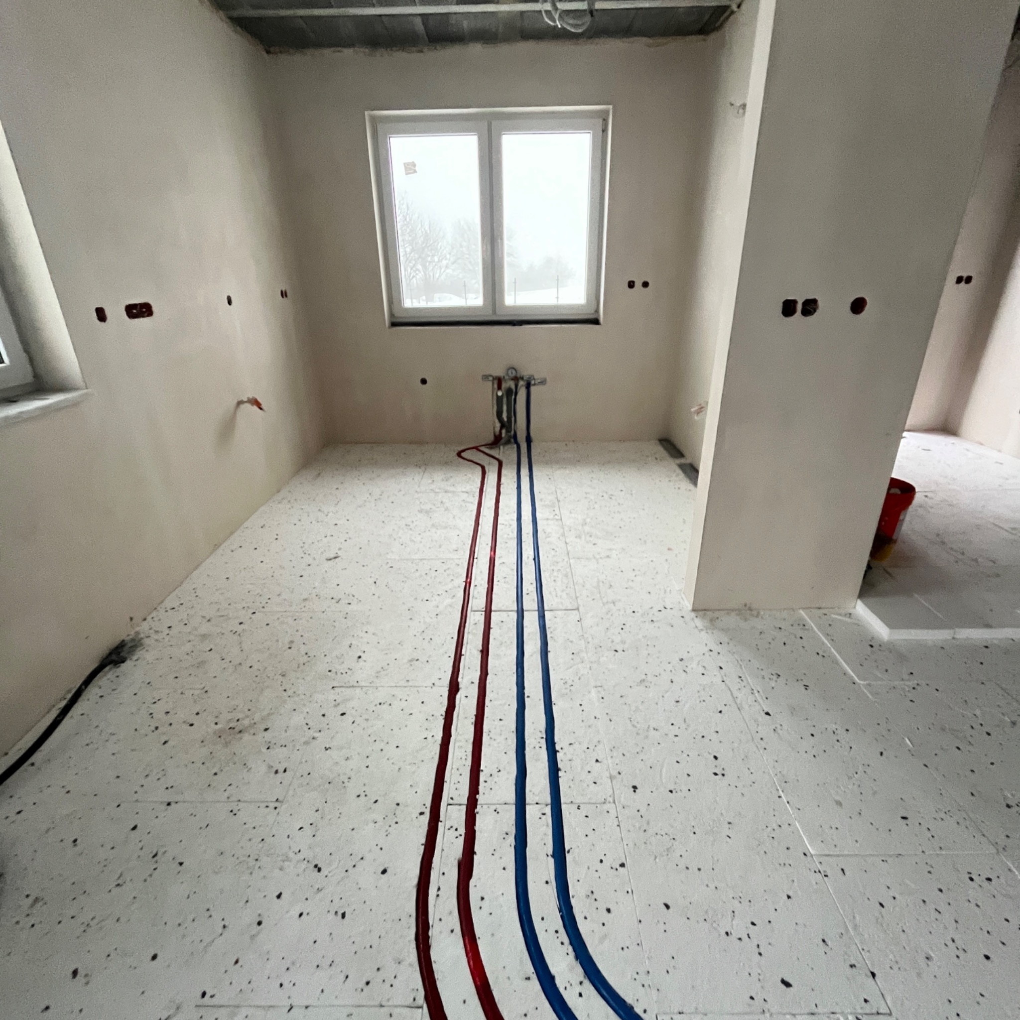 Wnętrze w trakcie prac hydraulicznych: czerwone i niebieskie rury prowadzone po podłodze z lastryko, przygotowane do podłączenia armatury pod oknem. Ściany w stanie surowym.