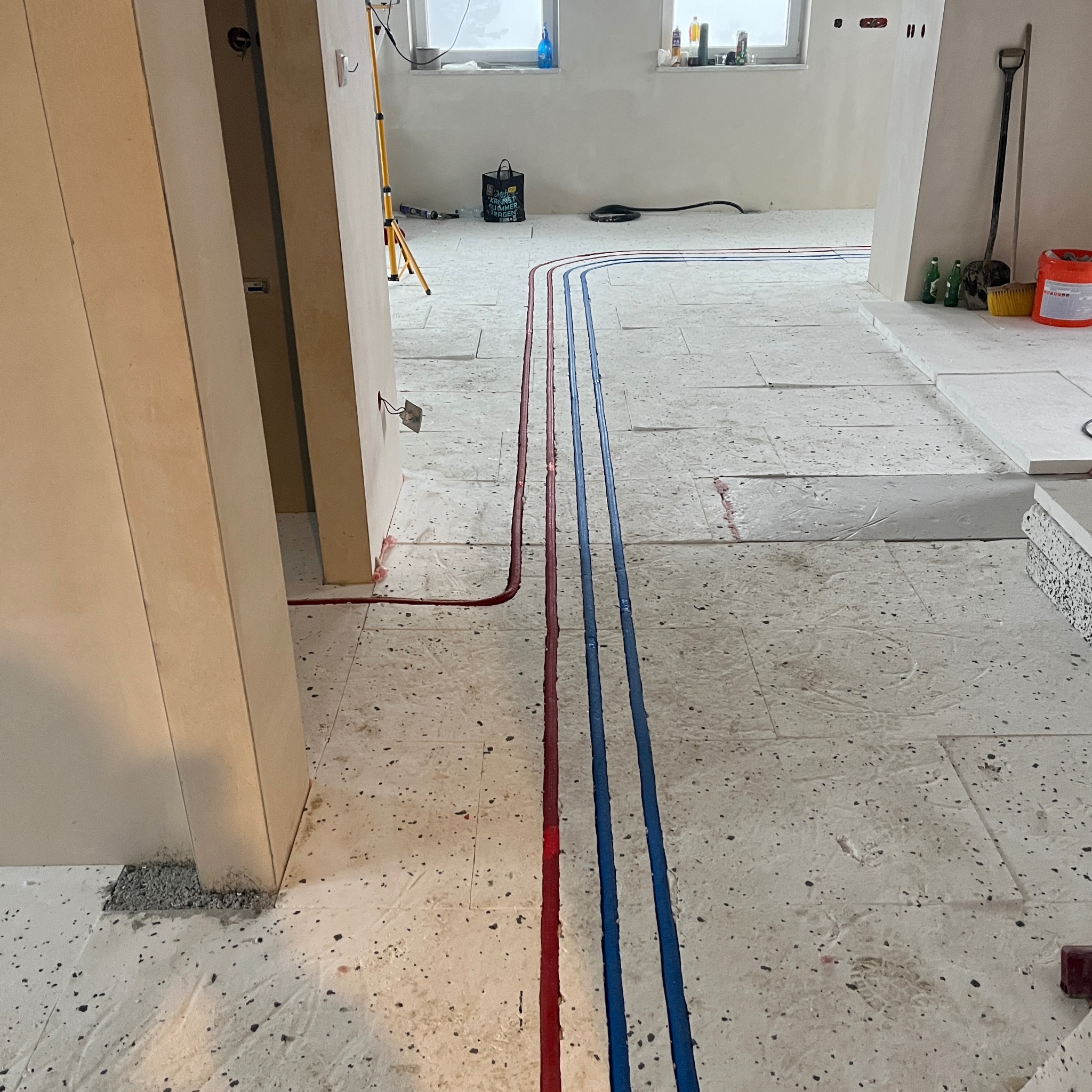 Ułożone rury ogrzewania podłogowego (czerwone i niebieskie) na izolacji, widok z góry, przygotowane do zalania posadzką. W tle narzędzia i materiały budowlane.