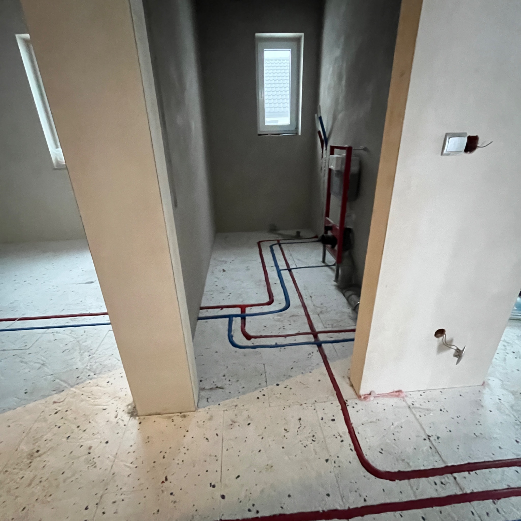 Wnętrze łazienki w trakcie remontu z widoczną instalacją wodną (czerwone i niebieskie rury) na podłodze i zamontowanym stelażem podtynkowym WC. Surowe ściany i okno.