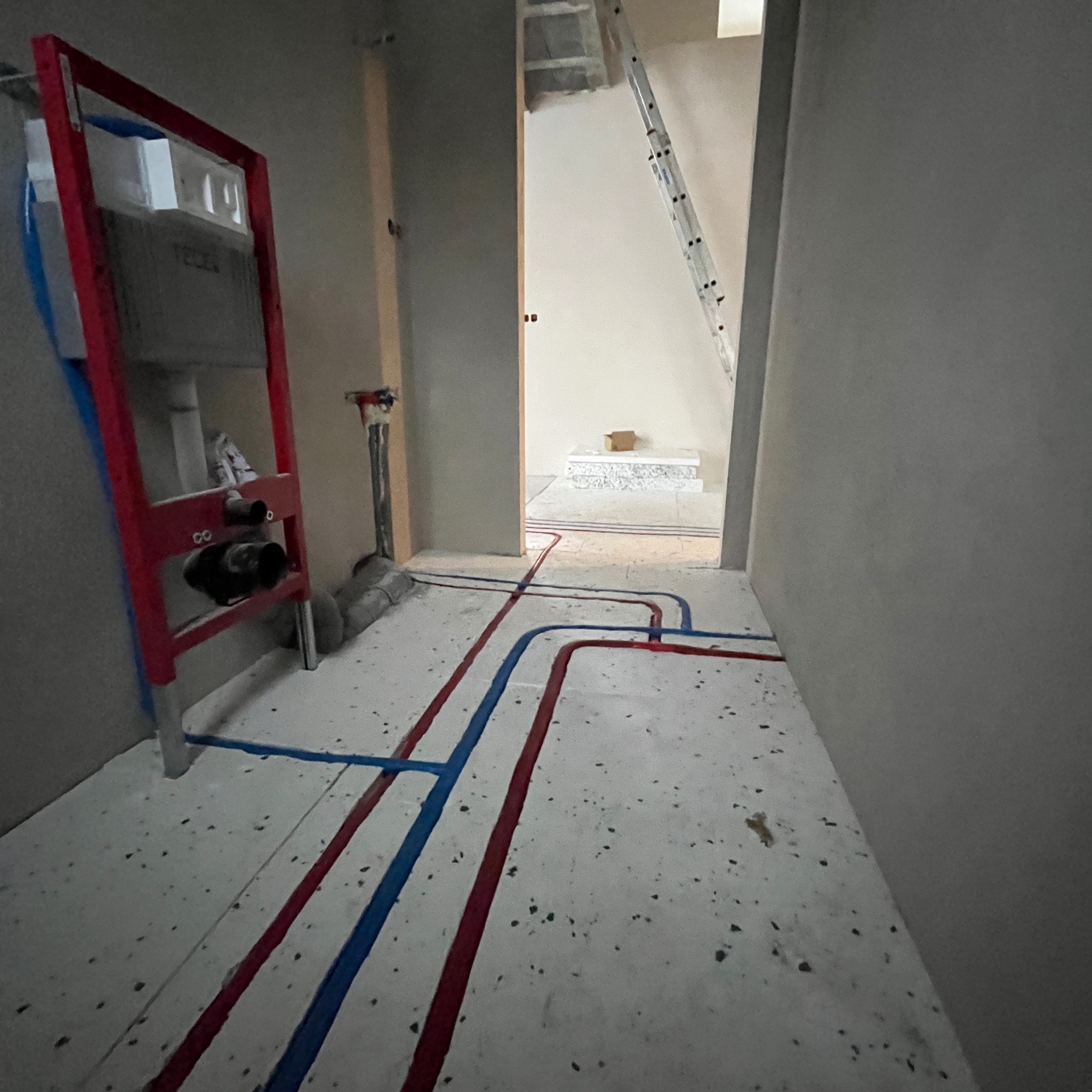 Instalacja podtynkowa WC z czerwono-niebieskimi rurami na podłodze w surowym stanie. W tle widoczna drabina i przygotowane podłoże pod płytki w kolejnym pomieszczeniu.