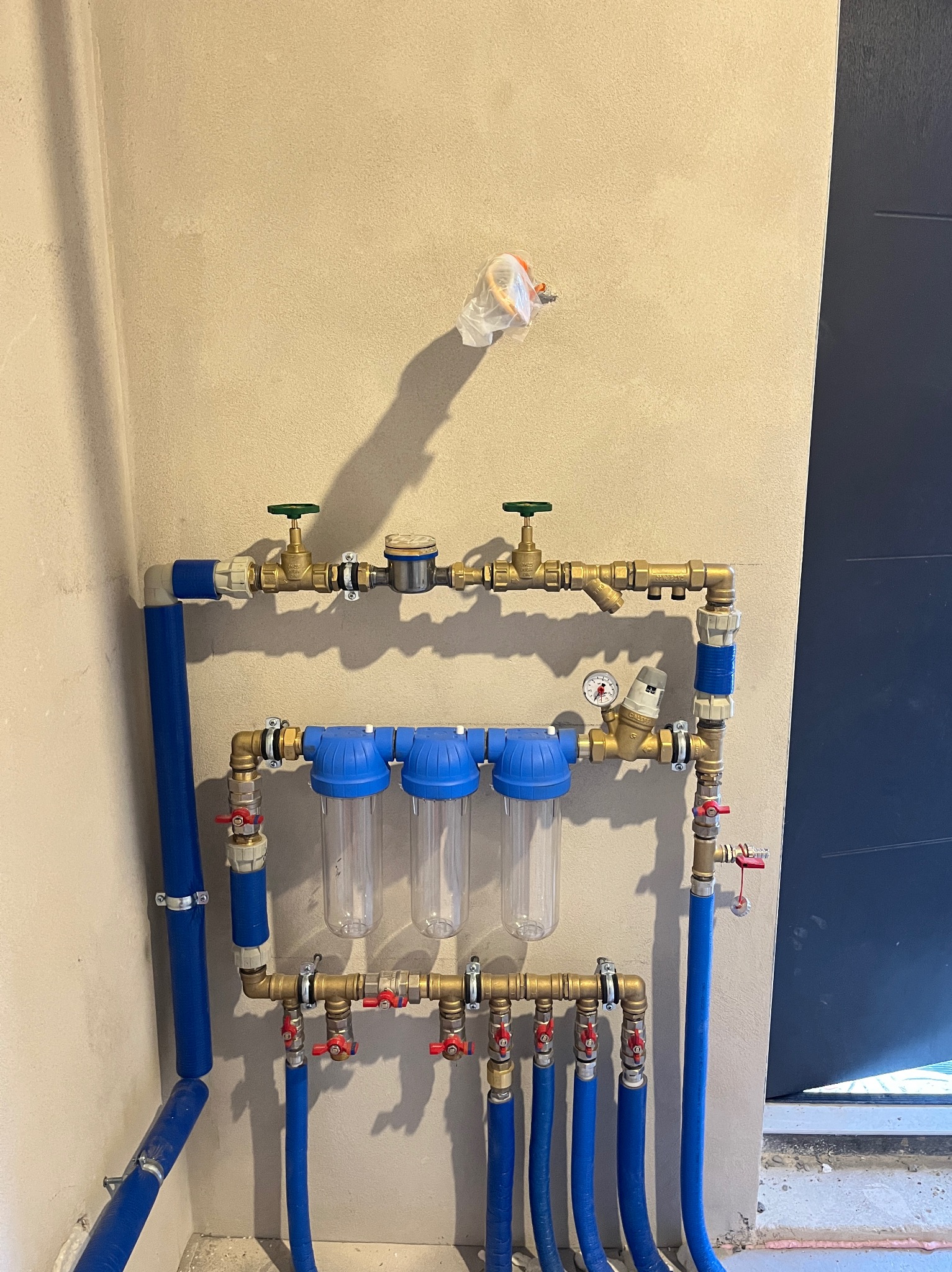 Złożona instalacja wodna z filtrami i zaworami, rury w kolorze niebieskim, mosiężne złączki, manometr. Widok na ścianę z instalacją, obok fragment drzwi.