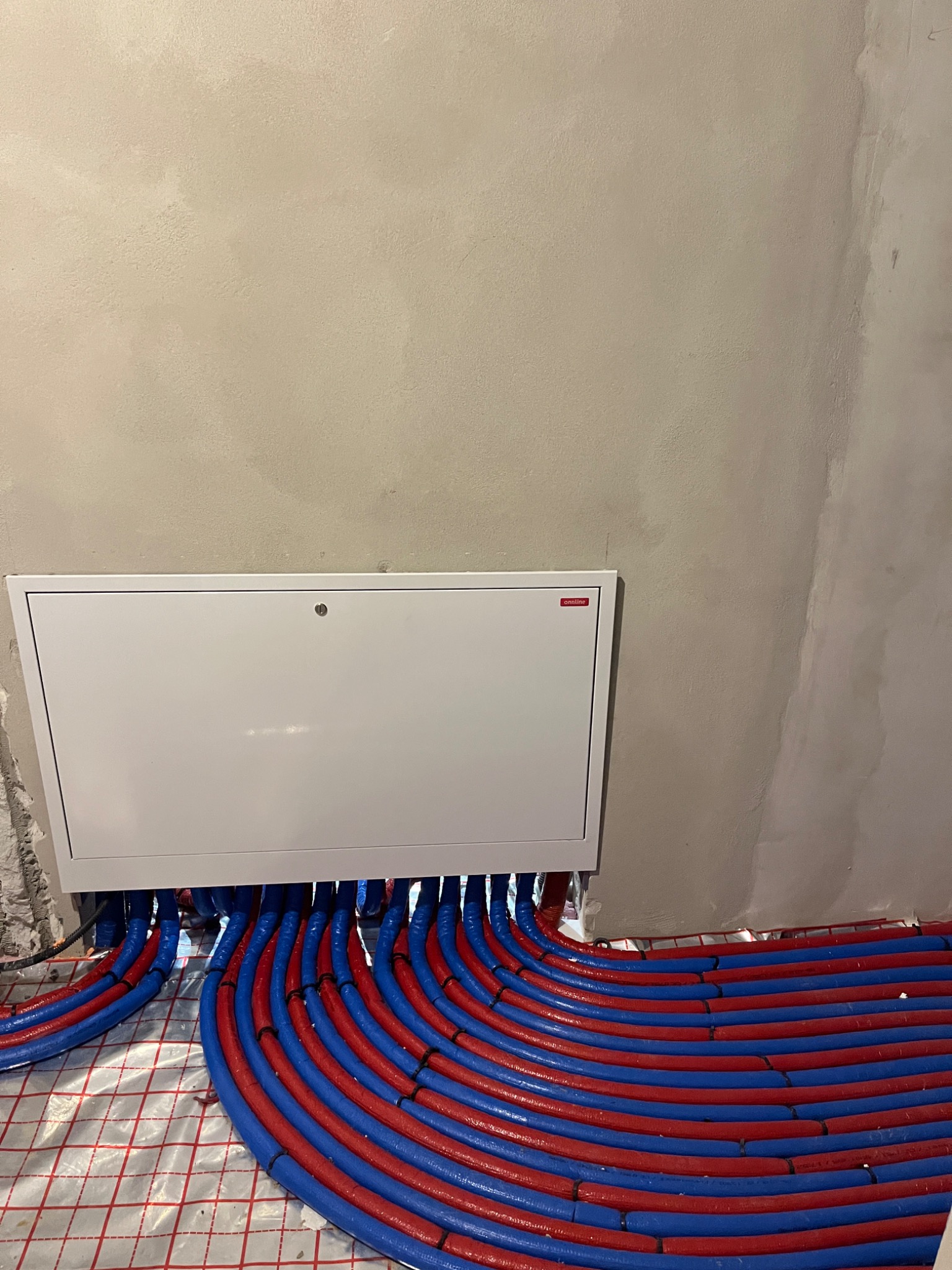 Rozłożone rury ogrzewania podłogowego w kolorze niebieskim i czerwonym, wpięte do białej skrzynki rozdzielczej na tle surowej ściany. Widoczna folia izolacyjna z rastrem.