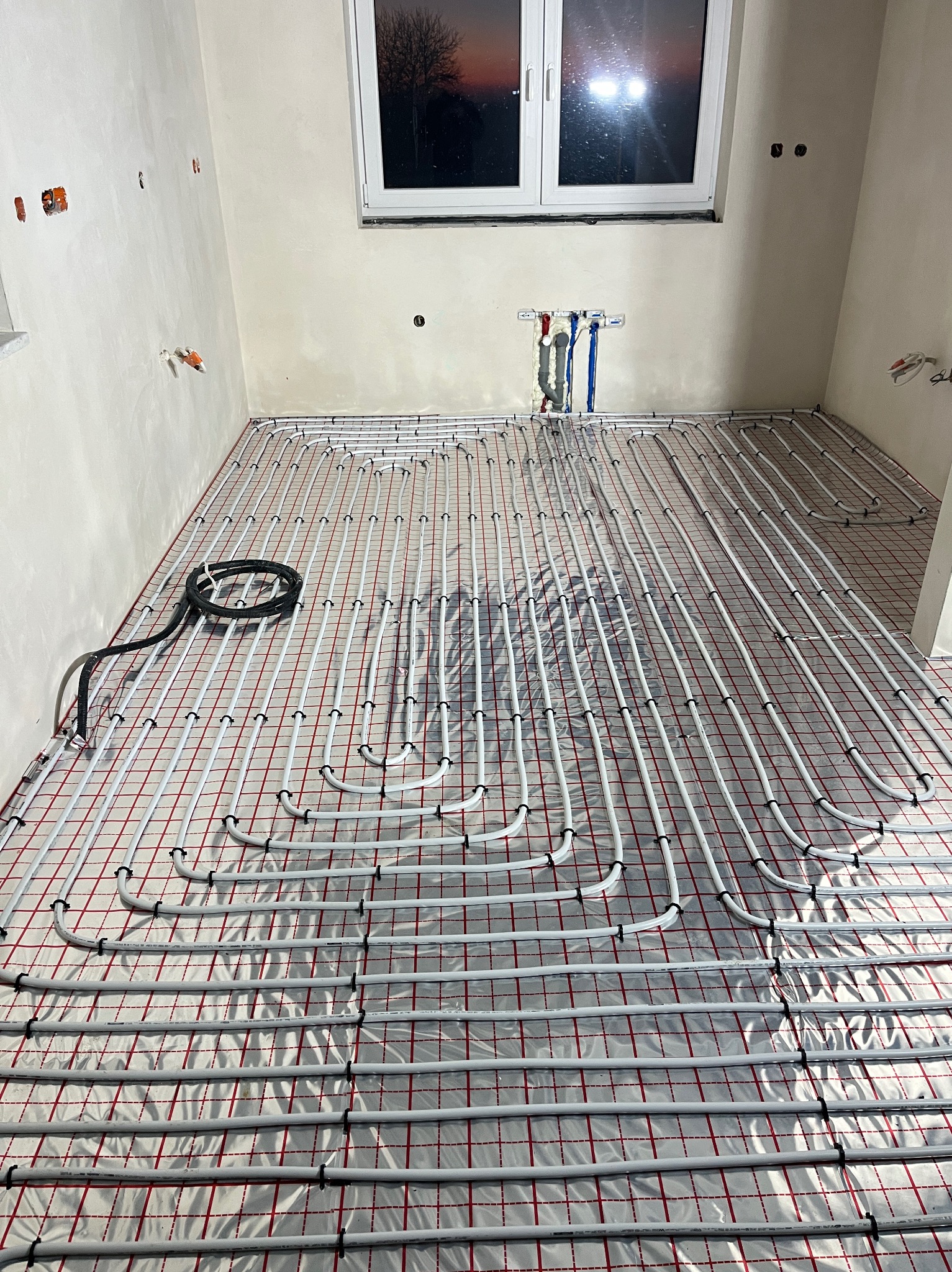 Białe rury ogrzewania podłogowego ułożone na folii z czerwoną siatką w surowym wnętrzu. Widoczne przyłącza do instalacji na ścianie i czarny przewód.