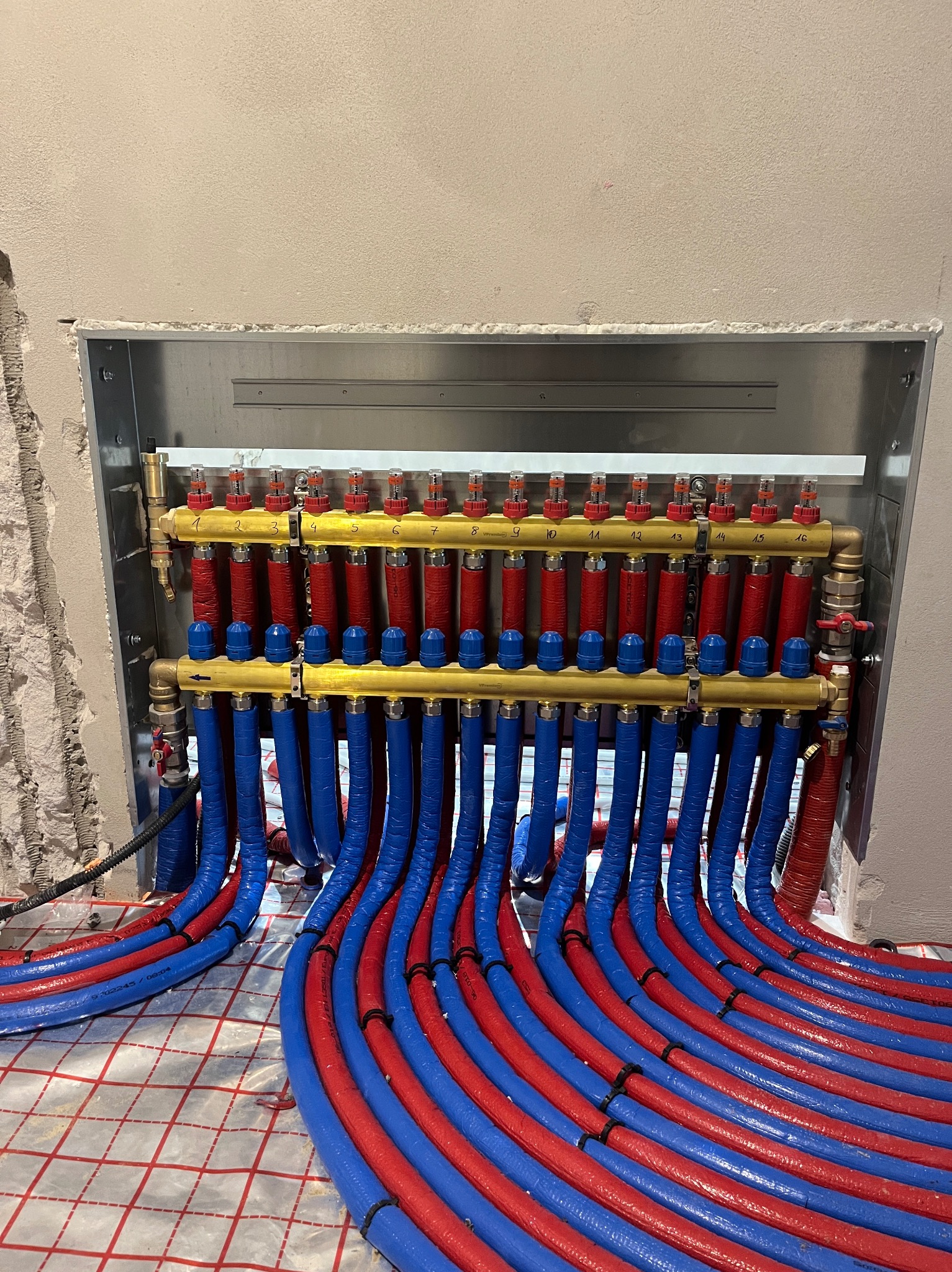 Rozdzielacz ogrzewania podłogowego z czerwono-niebieskimi rurami, zamontowany w metalowej obudowie w surowym stanie, widoczne fragmenty instalacji na podłodze.