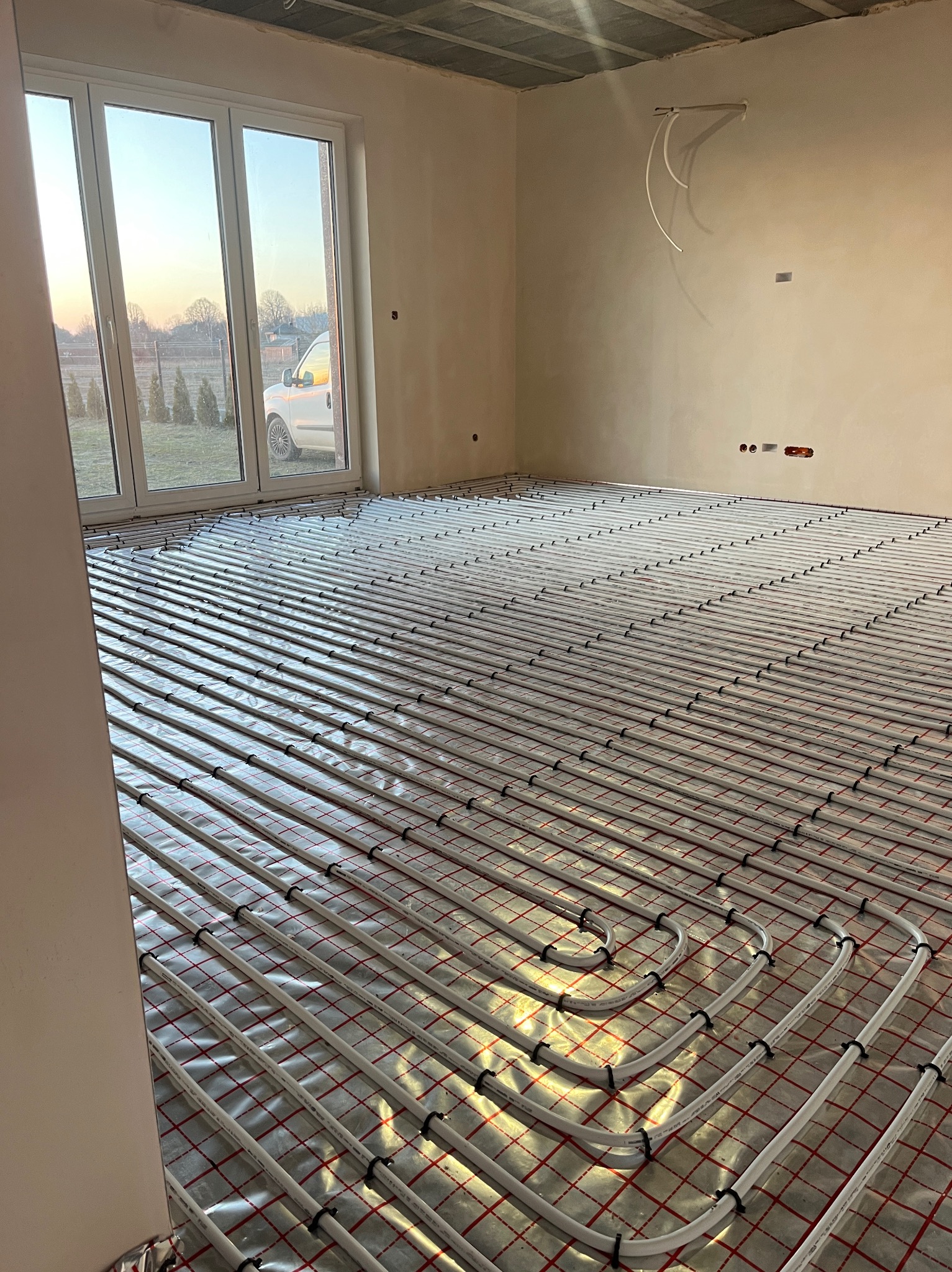 Białe rury ogrzewania podłogowego ułożone na folii w jasnym pokoju z oknem. Widoczne mocowania rur i fragment instalacji na ścianie. W tle dom za oknem.