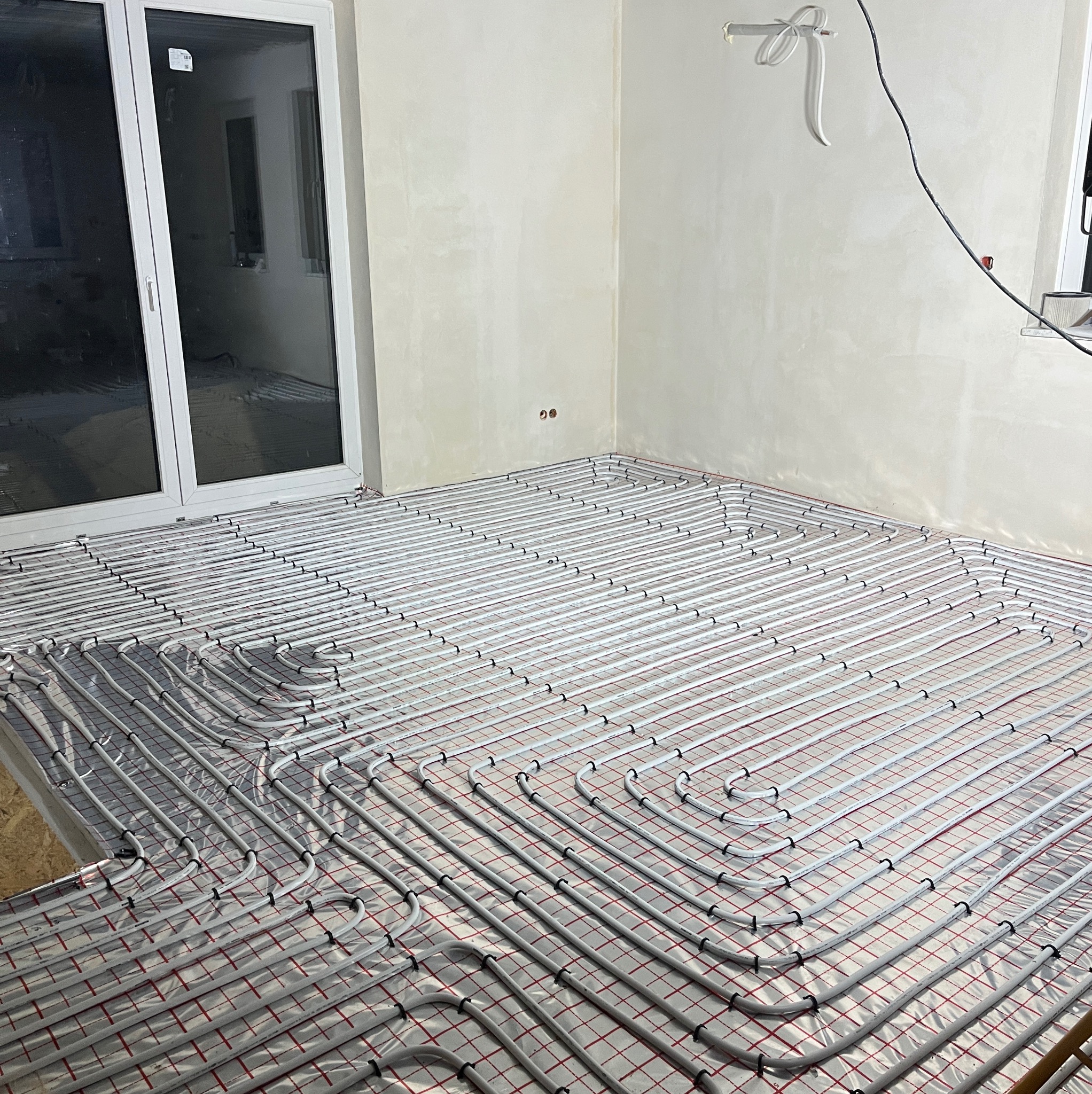 Białe rury ogrzewania podłogowego ułożone na folii z czerwoną siatką w jasnym pomieszczeniu z dużym oknem balkonowym. Widoczne elementy instalacji elektrycznej na ścianie.