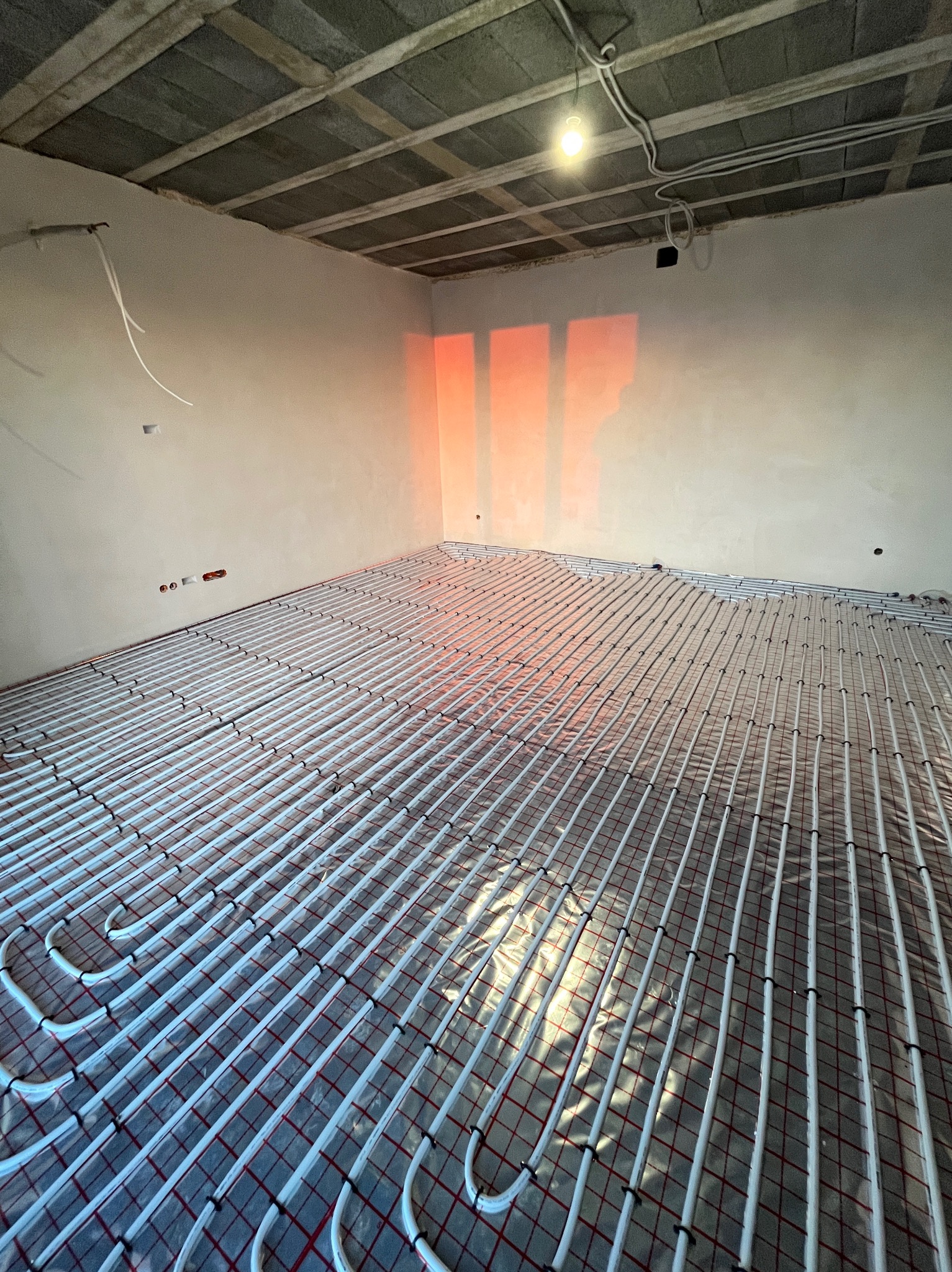 Białe rury ogrzewania podłogowego ułożone na folii w surowym wnętrzu. Na ścianie widoczne czerwone refleksy światła. Widok z góry na instalację przed wylaniem posadzki.
