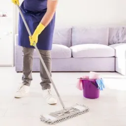 Osoba w niebieskim fartuchu i żółtych rękawicach mopuje białą podłogę w jasnym salonie z fioletową sofą; fioletowe wiadro z akcesoriami do sprzątania obok.