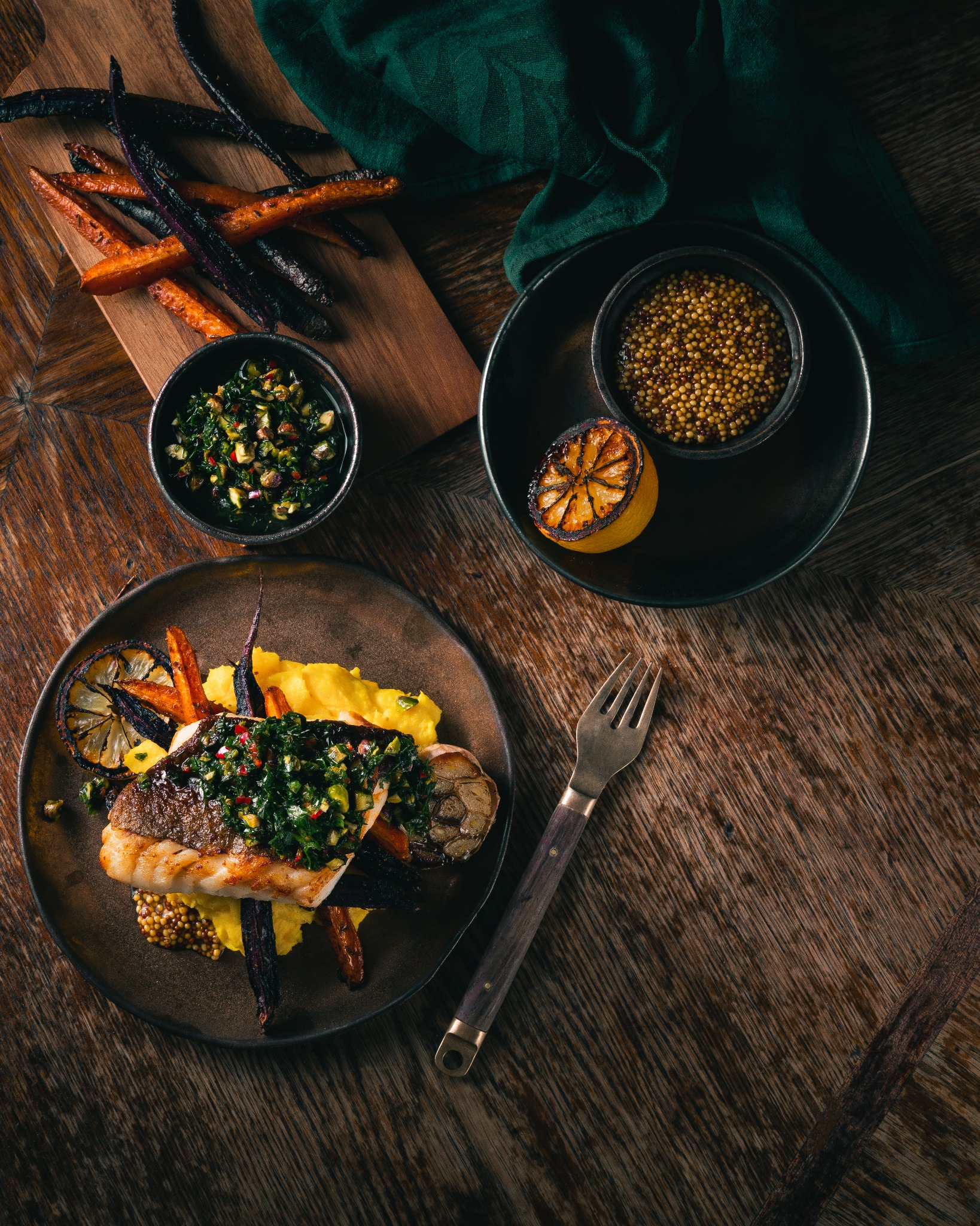 Elegancko podane danie: grillowana ryba z zielonym sosem chimichurri, puree, pieczone fioletowe i pomarańczowe marchewki, grillowana cytryna, musztarda ziarnista, serwowane na ciemnym talerzu...