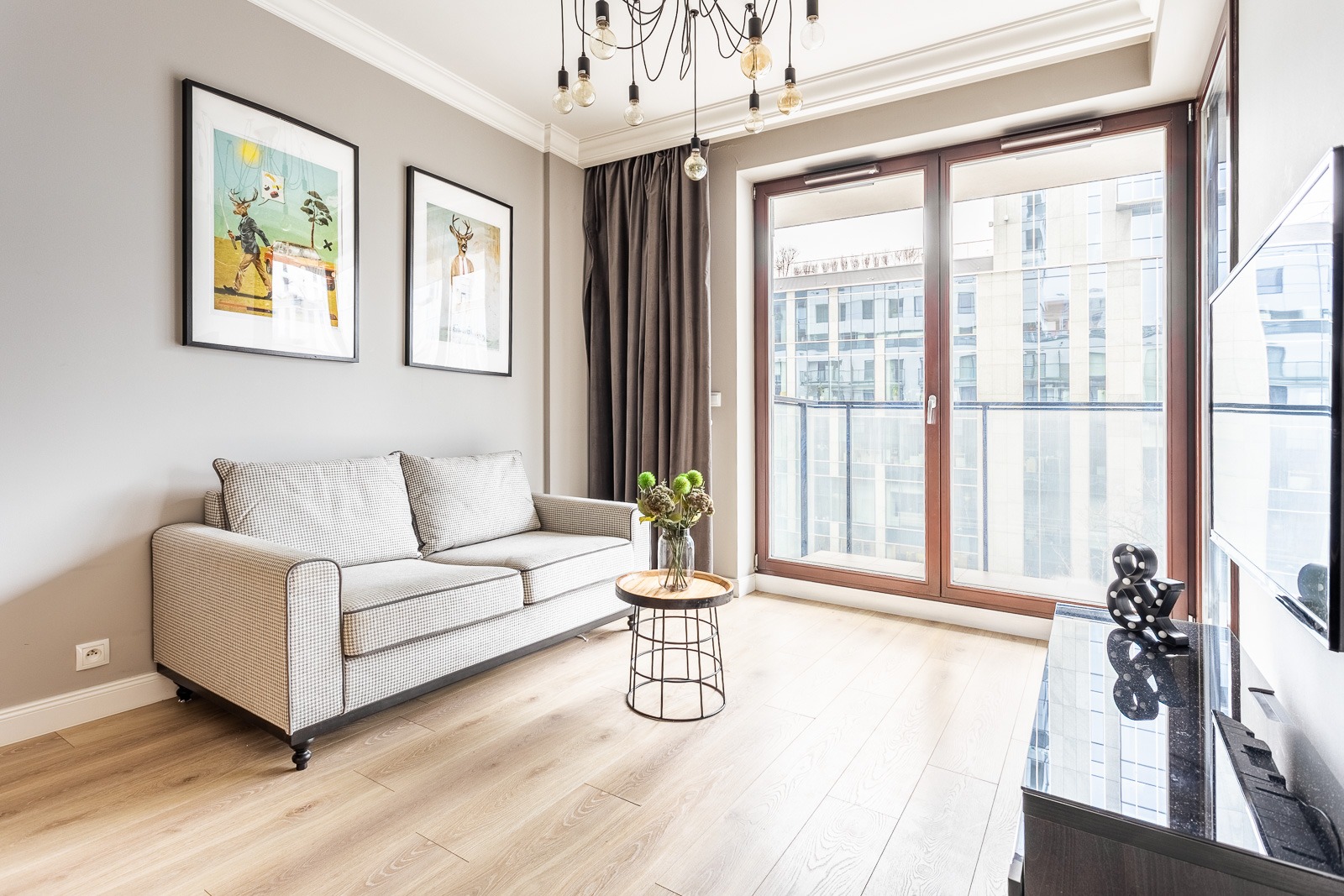 Jasny salon z szarą sofą w kratę, stolikiem z kwiatami i widokiem na nowoczesny budynek z balkonu.