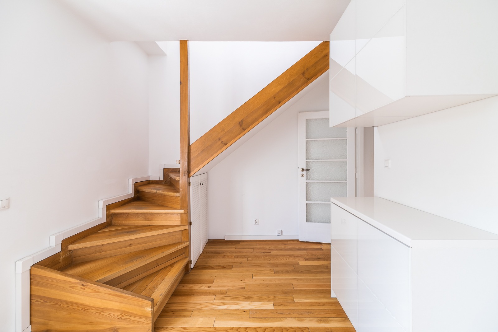 Wnętrze z drewnianymi schodami spiralnymi, białymi ścianami, drzwiami z szybkami i minimalistyczną zabudową kuchenną.