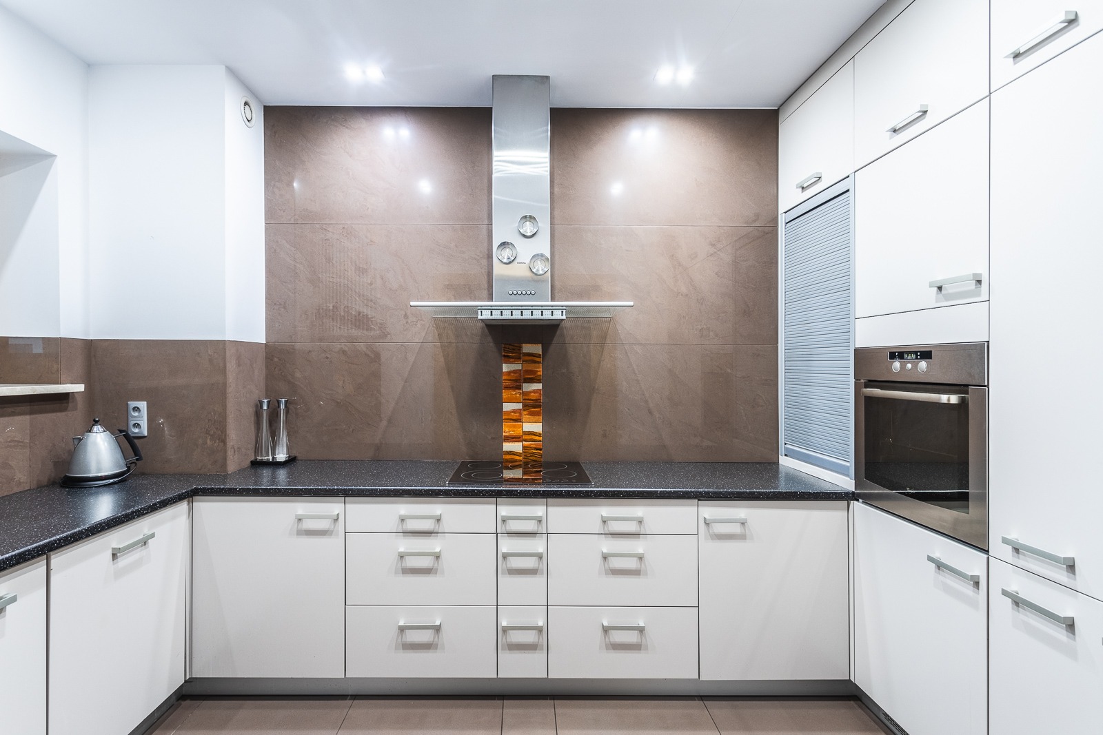 Nowoczesna, minimalistyczna kuchnia z białymi szafkami, czarnym blatem i brązowymi płytkami na ścianie. Stalowy okap z dekoracyjnym elementem z mozaiki nad płytą indukcyjną. Widoczny czajnik...