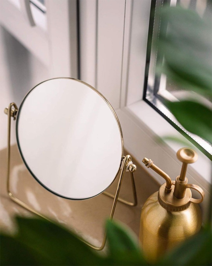 Okrągłe lusterko kosmetyczne w złotej ramie i ozdobny, mosiężny spryskiwacz do roślin na parapecie okna w Warszawie, otoczone zielenią.