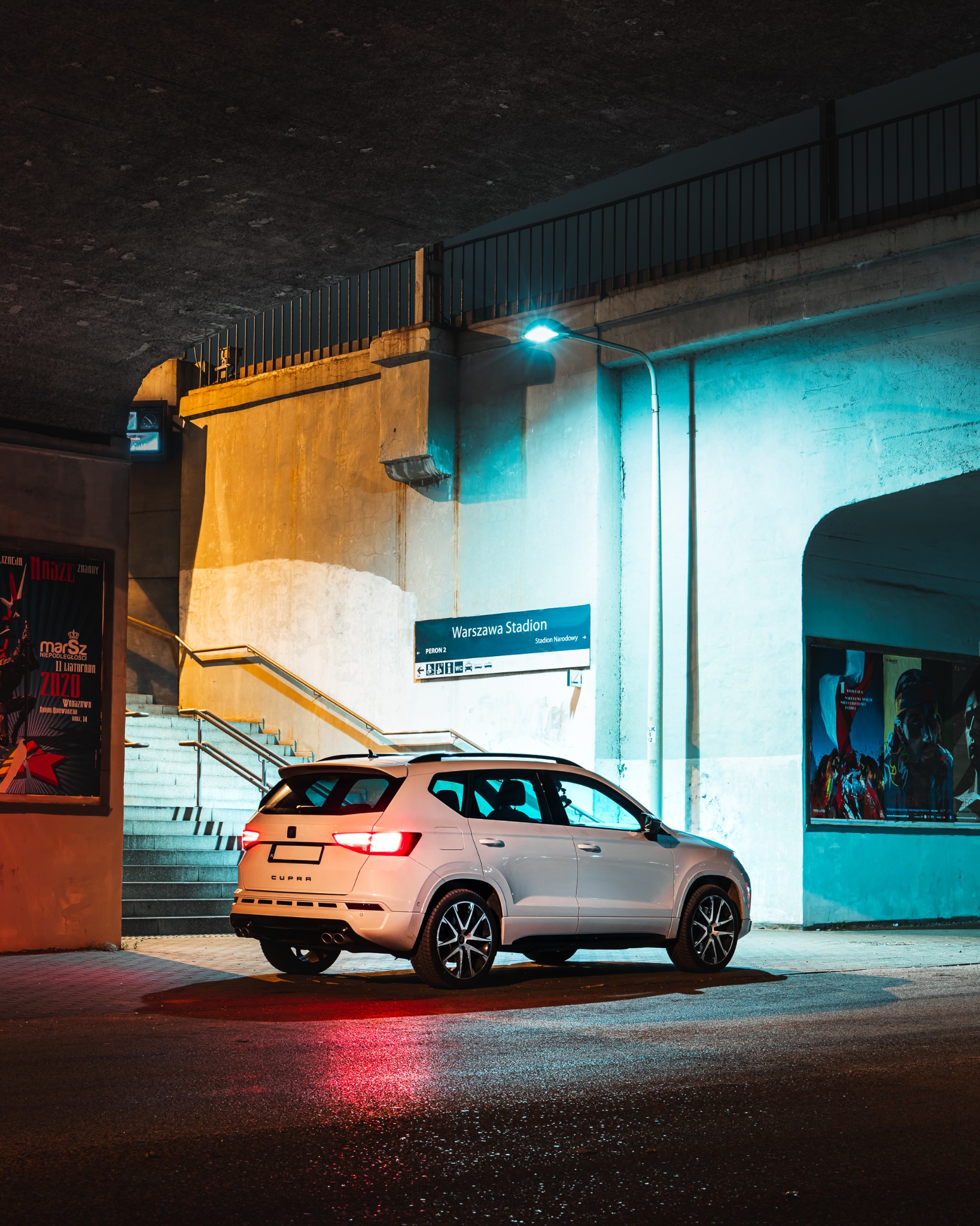 Biały samochód Cupra stoi nocą pod wiaduktem w pobliżu stacji Warszawa Stadion, światła stopu odbijają się na mokrym asfalcie.
