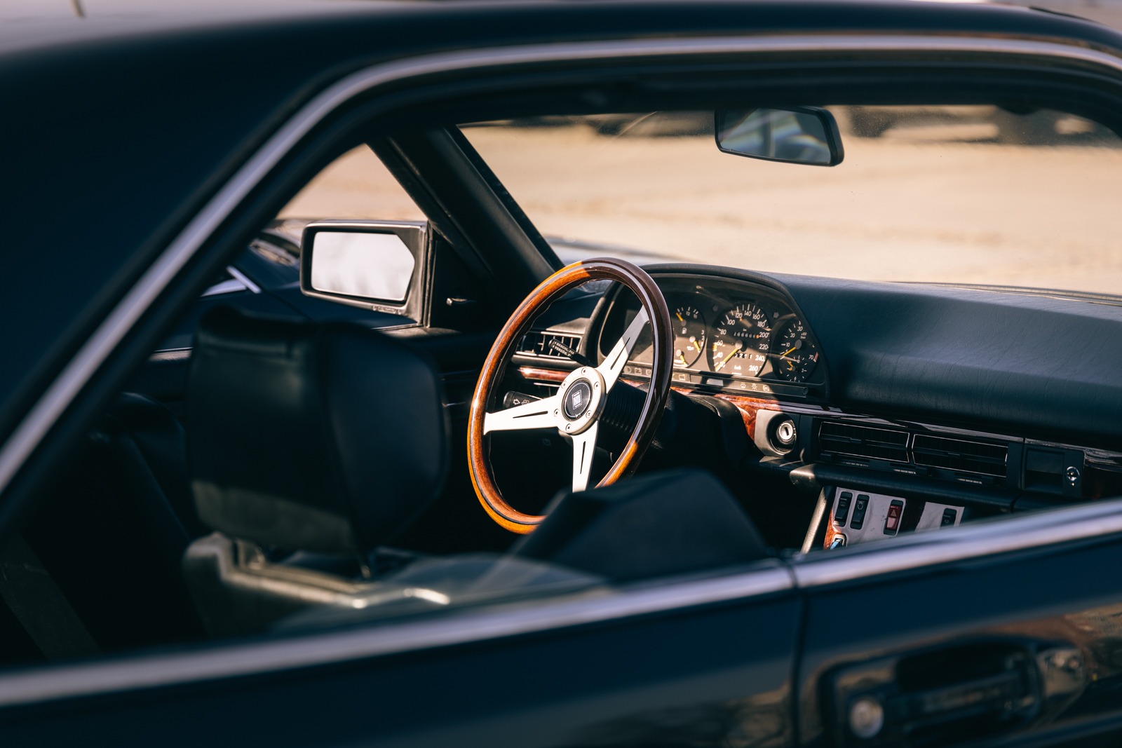 Wnętrze klasycznego samochodu z drewnianą kierownicą i czarną skórzaną tapicerką, widoczne przez otwarte okno.