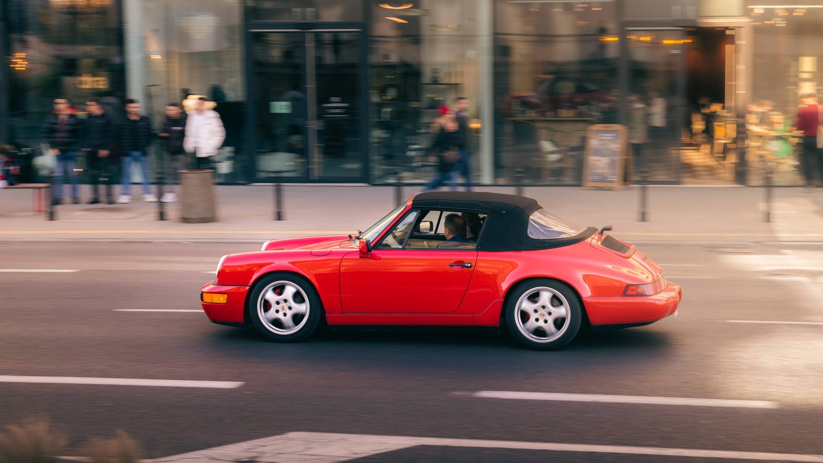 Dynamiczne ujęcie czerwonego kabrioletu Porsche 911 jadącego ulicą miasta, z rozmytymi postaciami w tle i odbiciami w szybach wystawowych.
