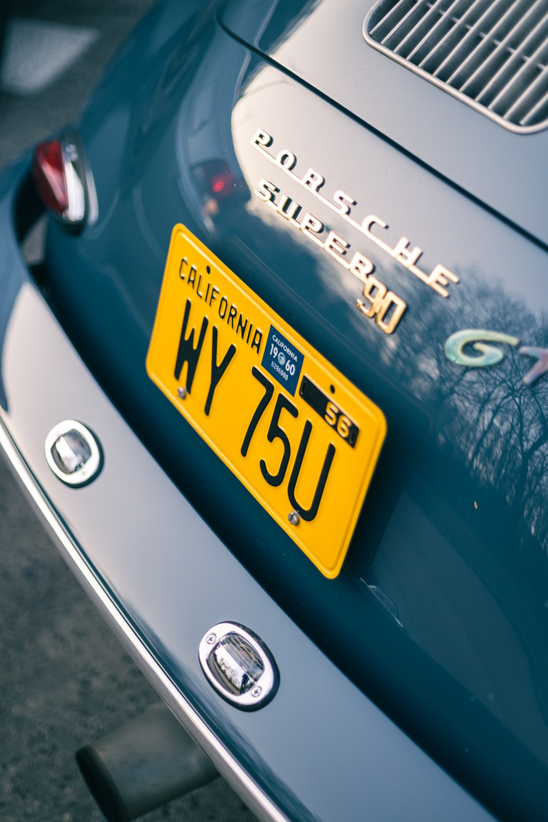Detale tylnej części klasycznego samochodu Porsche Super 90 GT z żółtą kalifornijską tablicą rejestracyjną WY 75U, rok 1960, widoczne chromowane elementy i odblask drzew na lakierze.