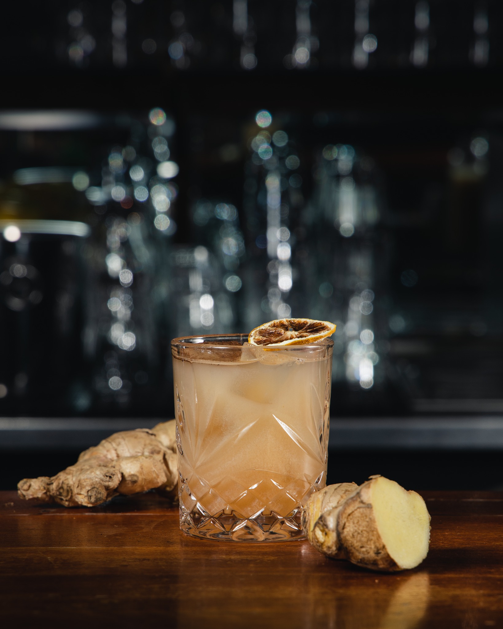 Kryształowa szklanka z bursztynowym napojem i lodem, udekorowana suszoną pomarańczą, obok korzenie imbiru na drewnianym blacie, tło rozmyte.